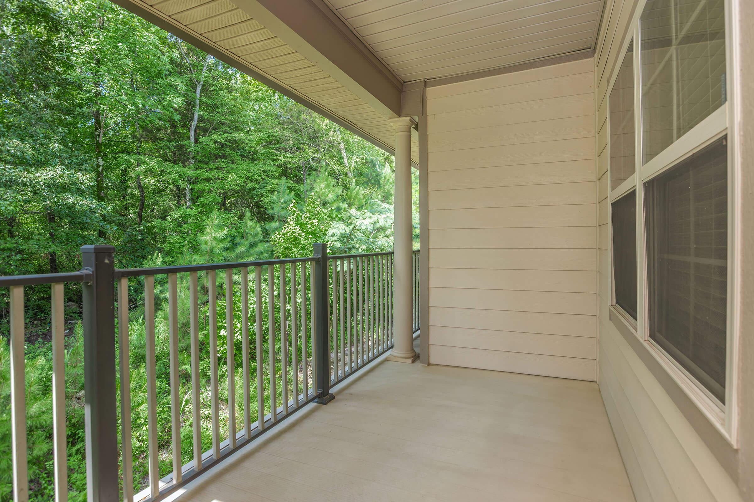 Empty balcony with a railing, overlooking lush green trees and foliage. The space has beige walls and is well-lit, providing a serene outdoor connection.