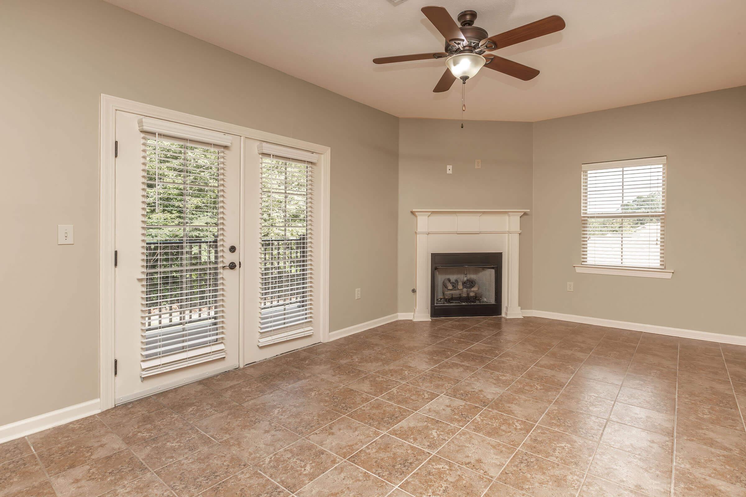 Interior view of a spacious living room featuring a ceiling fan, a corner fireplace, and large windows with blinds. The floor is tiled, and there are double doors leading to a balcony or outdoor space, creating a bright and inviting atmosphere.