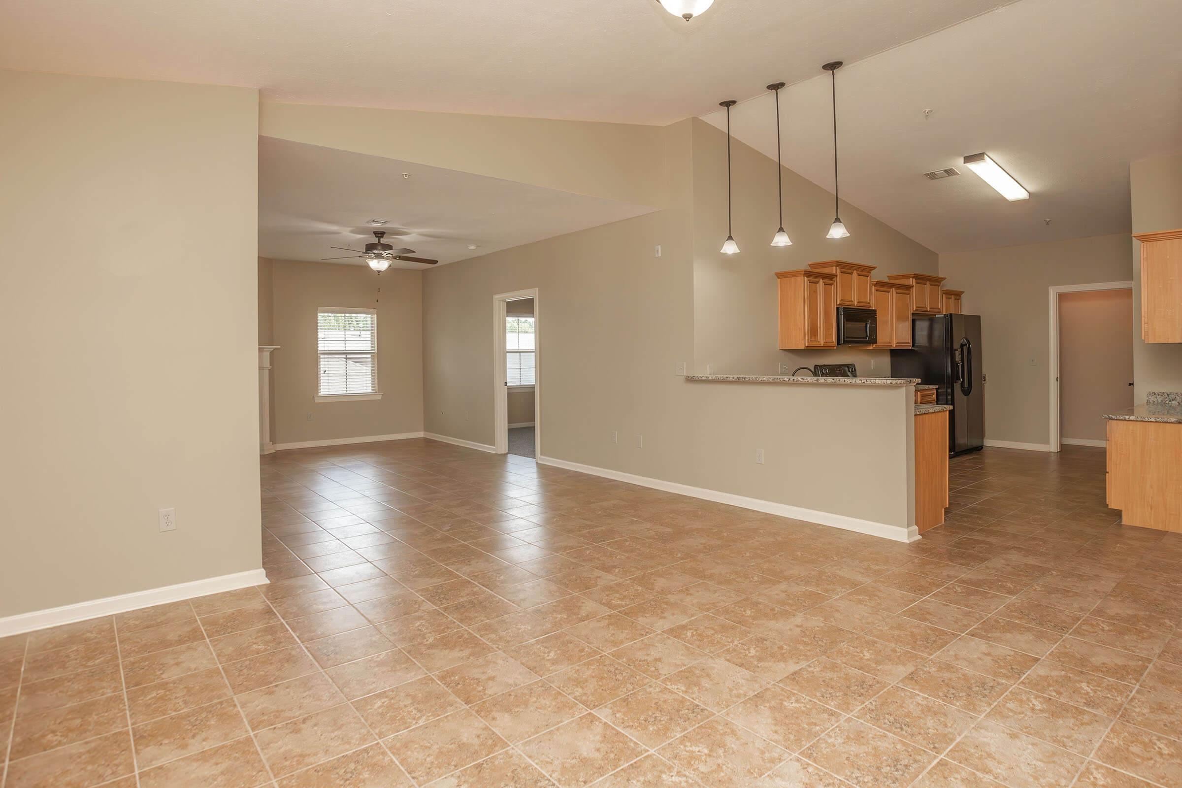 Spacious interior of a modern open-concept living area featuring tiled flooring, a kitchen with wooden cabinetry and black appliances, and large windows providing natural light. The room includes a ceiling fan and is designed for a comfortable, inviting atmosphere.