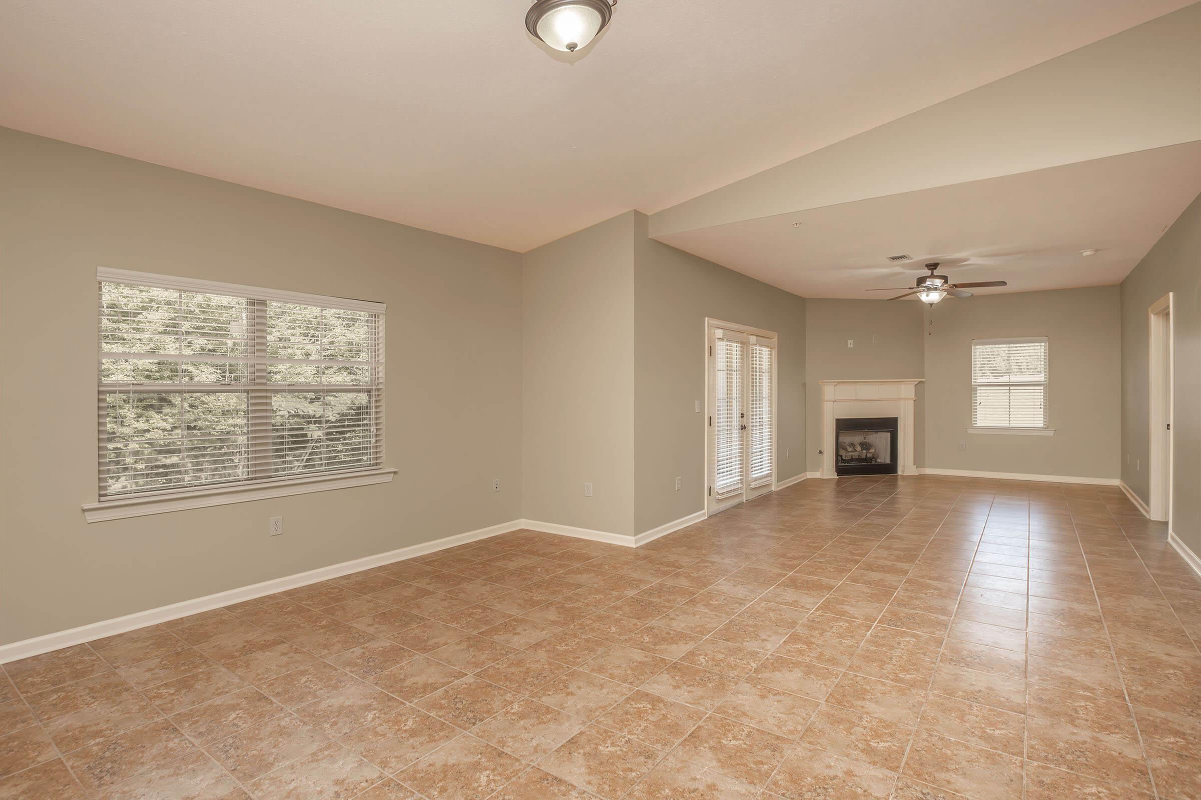 Spacious living room with light beige walls and tile flooring. Features large windows providing natural light and a ceiling fan. An empty fireplace on one side adds a focal point to the room. Double doors lead outside, creating an open and airy feel in the area.