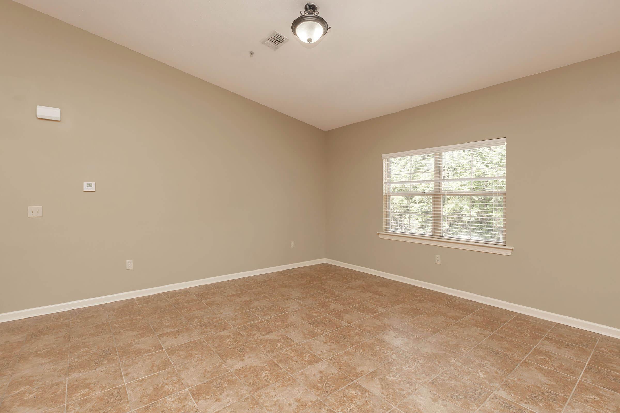 A spacious room with beige walls and tiled flooring. It features a single window with blinds, allowing natural light to enter. A ceiling light fixture is visible, and there are minimal decorative elements or furniture, creating an open and airy atmosphere.