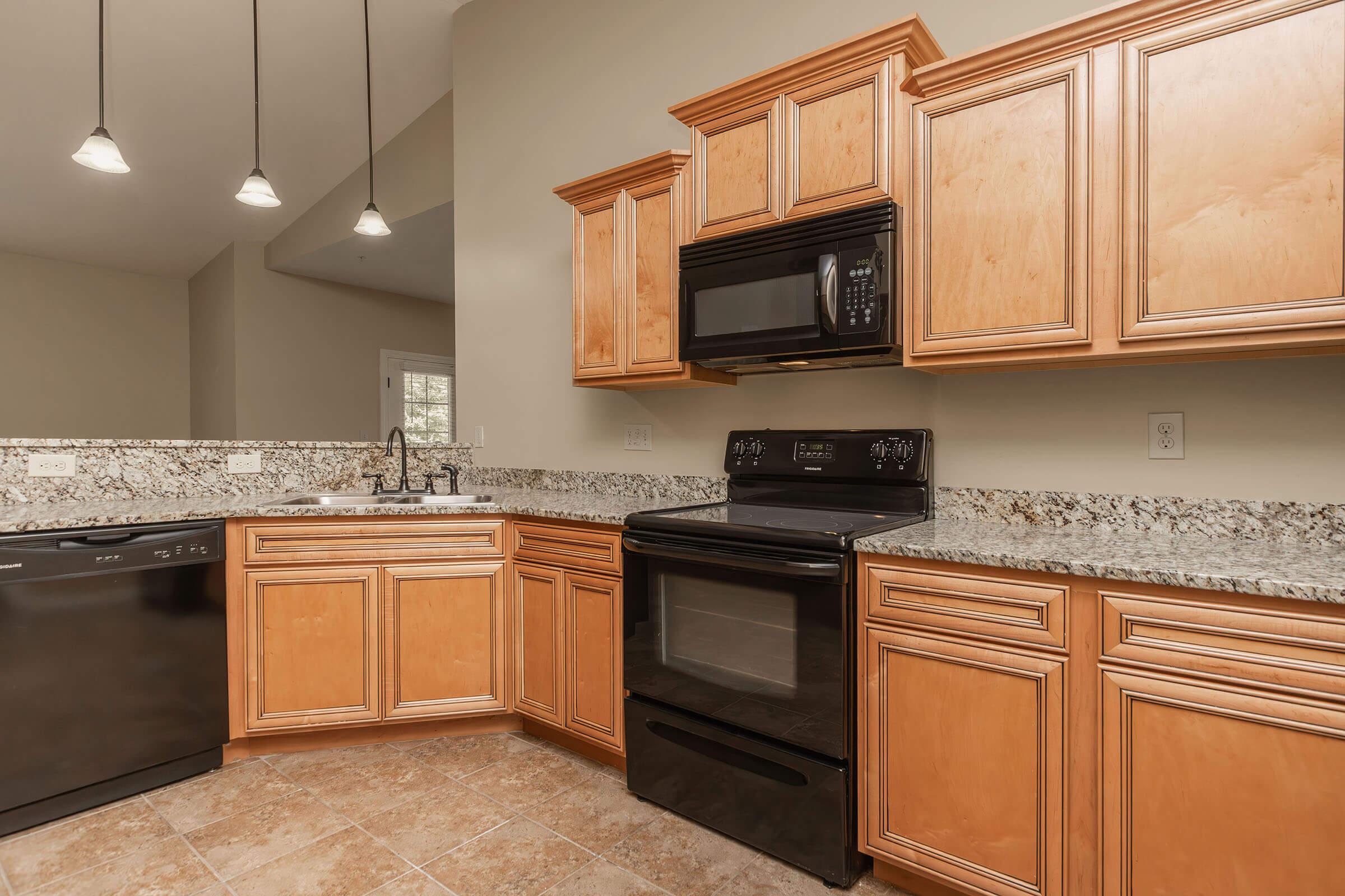 A modern kitchen featuring wooden cabinets, a black microwave above a black stove, and a black dishwasher. The countertop is adorned with granite, and there are pendant lights hanging from the ceiling, providing a bright and welcoming atmosphere. The floor is tiled in a neutral color.