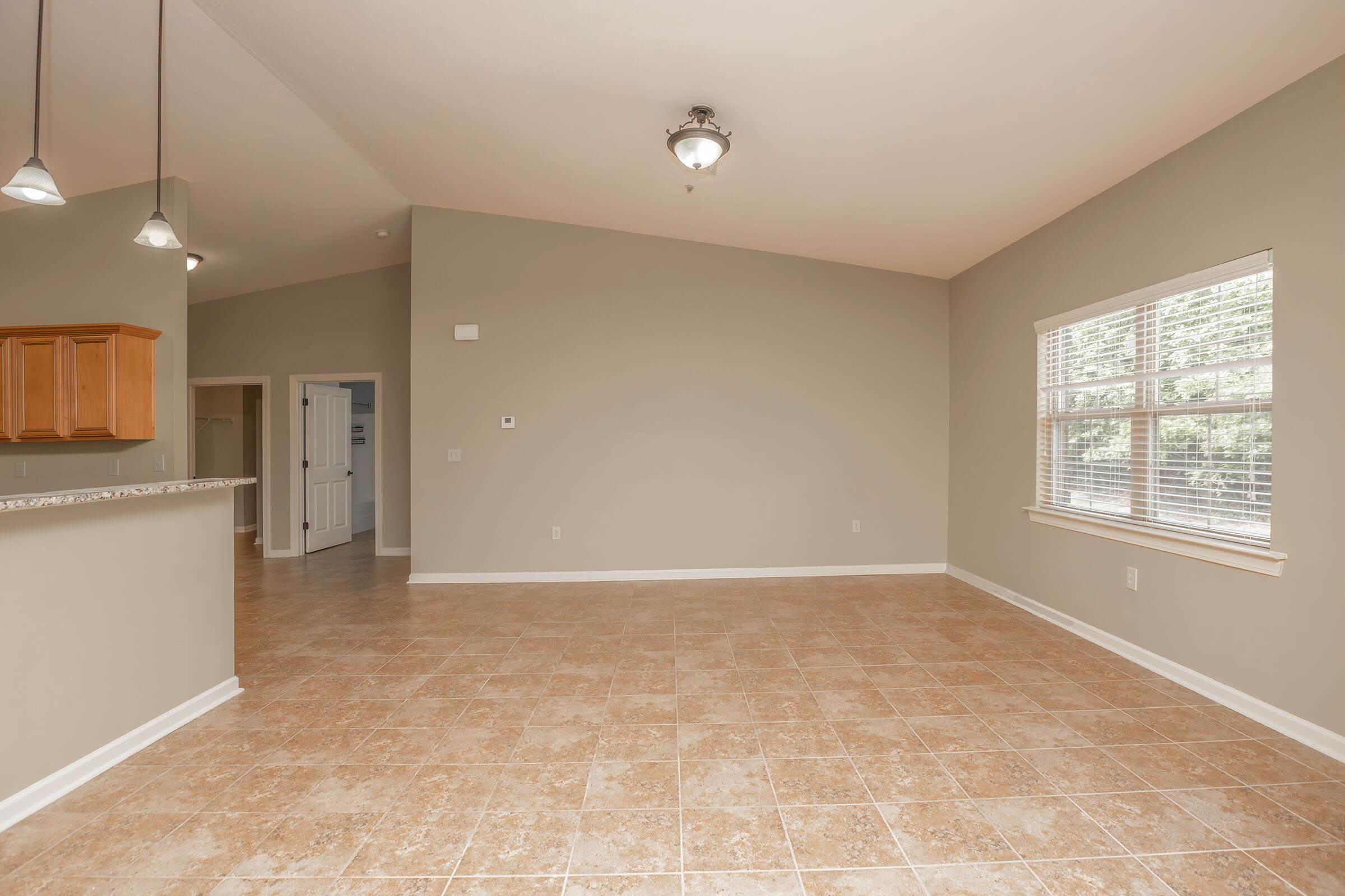 A spacious, empty living area with beige tiled flooring and light gray walls. There is a window allowing natural light, and a doorway leading to another room. The kitchen area is partially visible, featuring wooden cabinets and pendant lighting above.