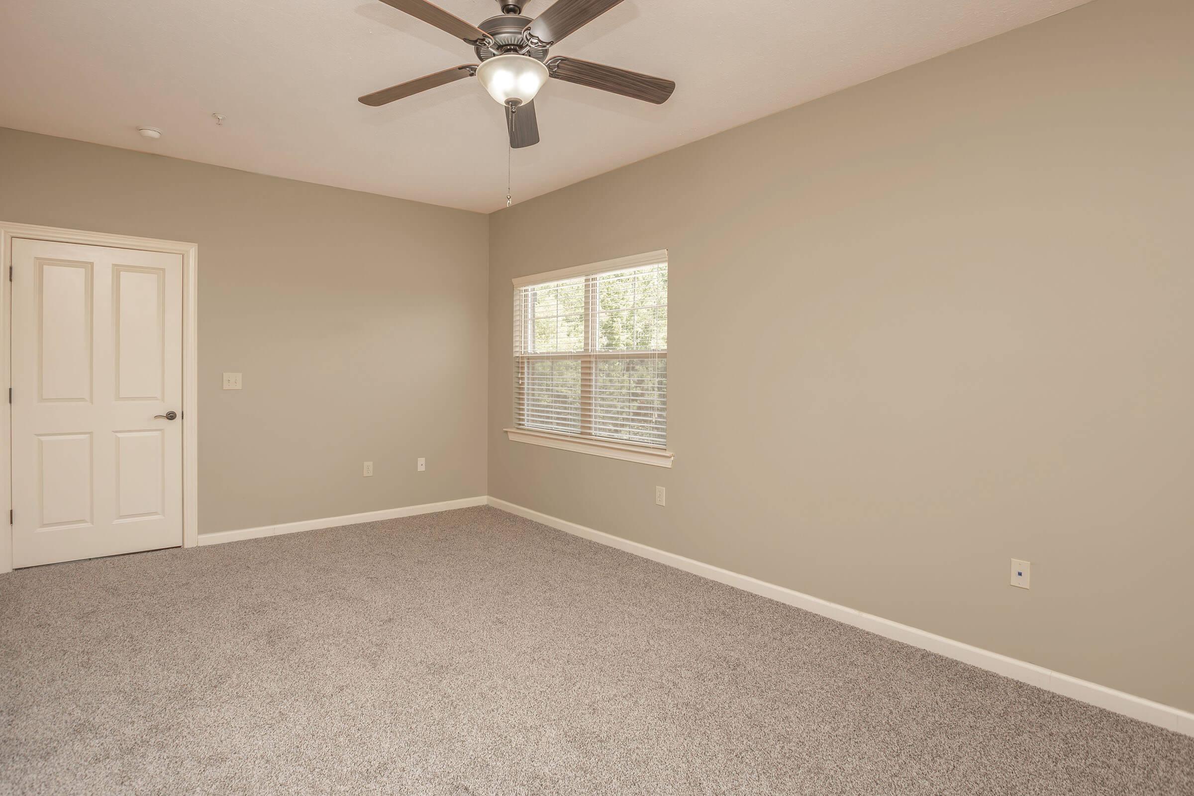A spacious, empty room with light beige walls and soft gray carpet. There is a ceiling fan with a light fixture, and a window allowing natural light to enter. A closed door is visible on one wall, contributing to the open and minimalistic feel of the space.
