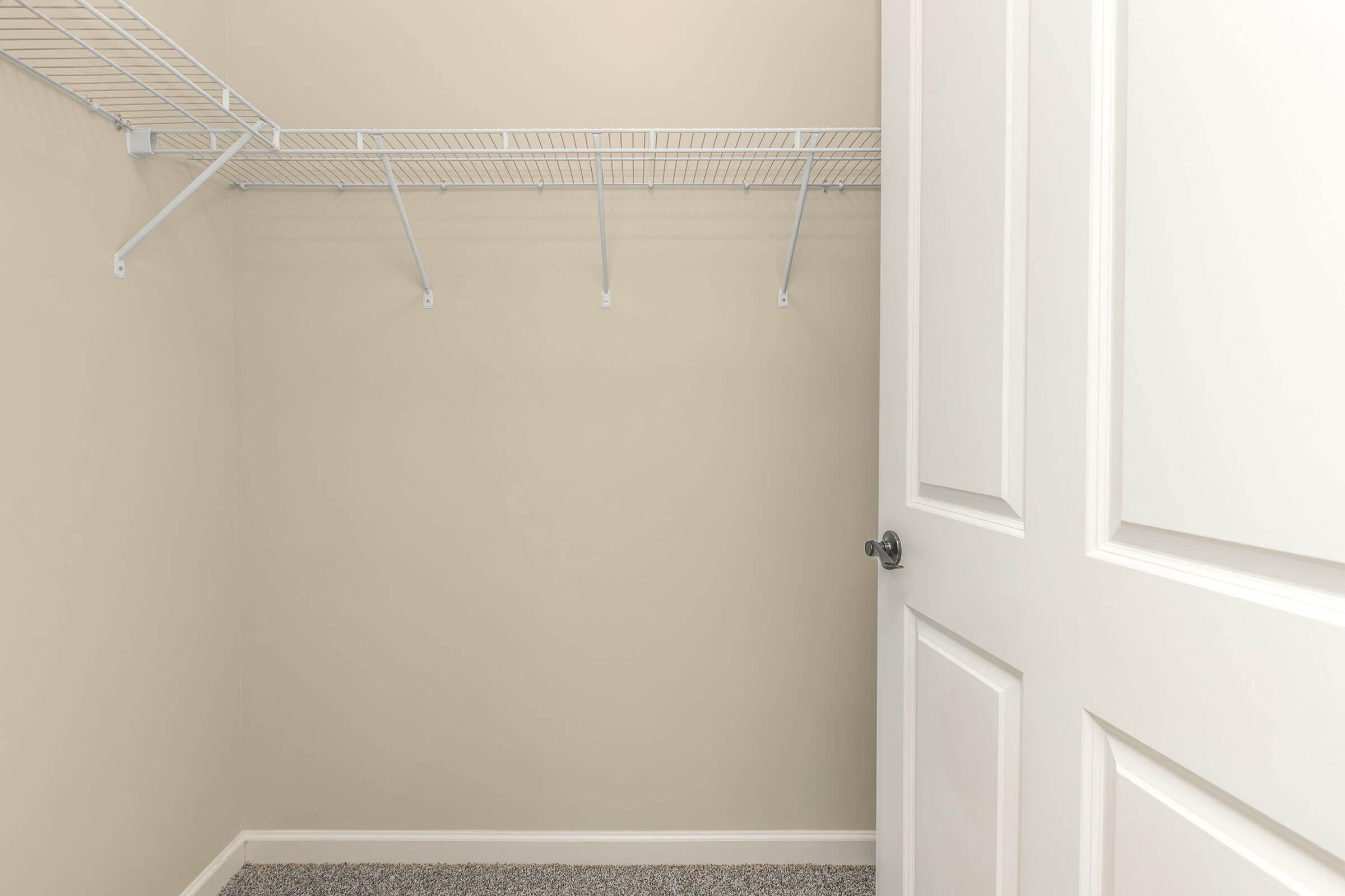 A view of an empty closet with beige walls and a wire shelf installed. The door is partially open, showing the interior space, which has no items or clothing hanging. The floor is carpeted in a neutral tone, complementing the simple design of the closet.