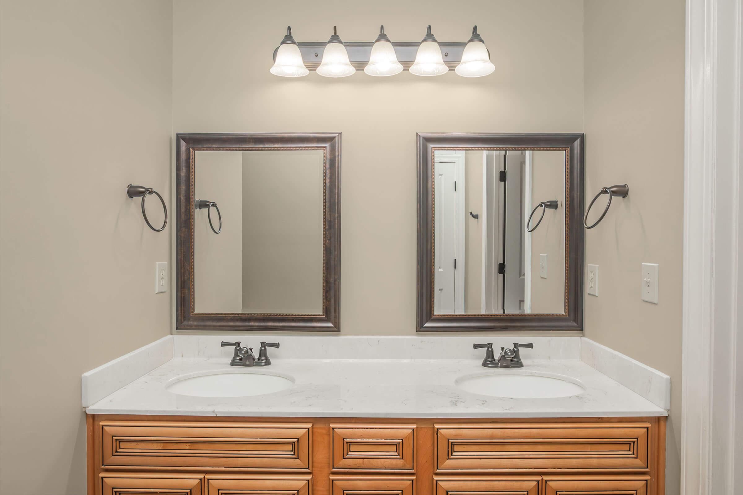 A well-lit bathroom featuring dual mirrors above a double sink vanity with wooden cabinetry. The wall-mounted light fixture consists of four bulbs, illuminating the space. The neutral-colored walls provide a calm backdrop, and the layout is symmetrical, showcasing clean lines and a modern design.