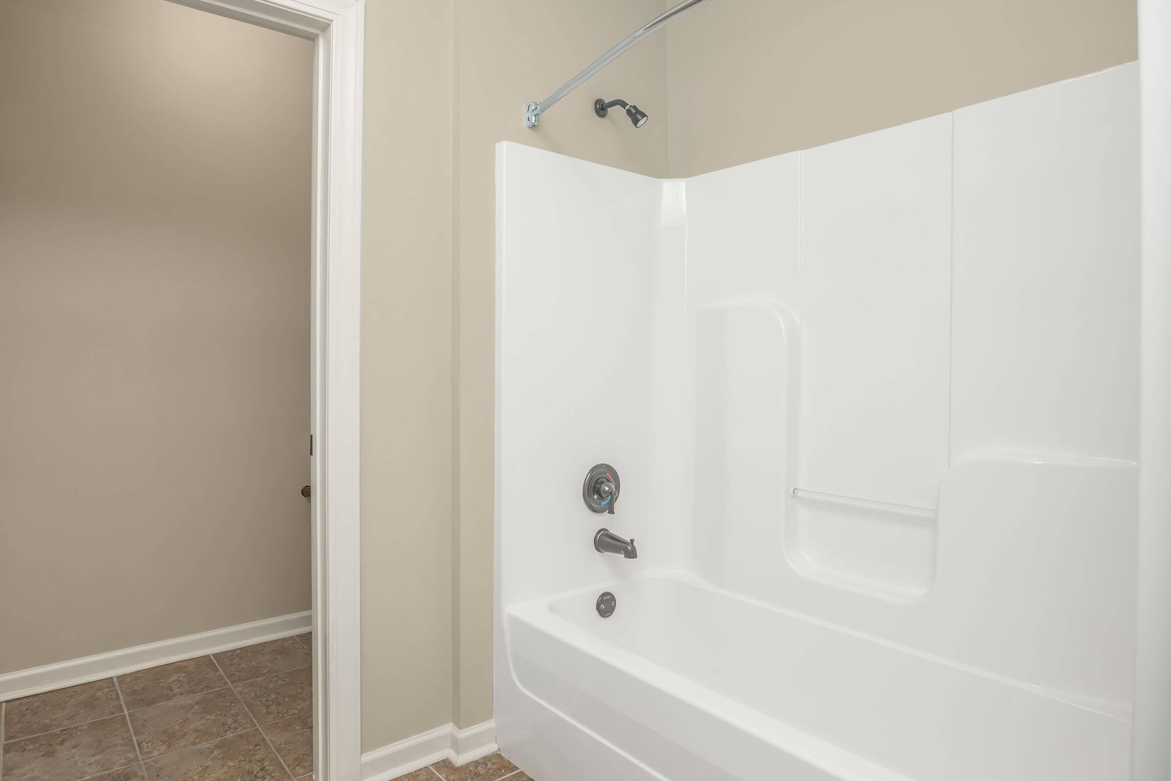 A clean, modern bathroom featuring a white shower tub combo with a chrome faucet and showerhead. The walls are painted a neutral tan color, and there is a doorway leading to another room on the left. The floor has light-colored tiles.