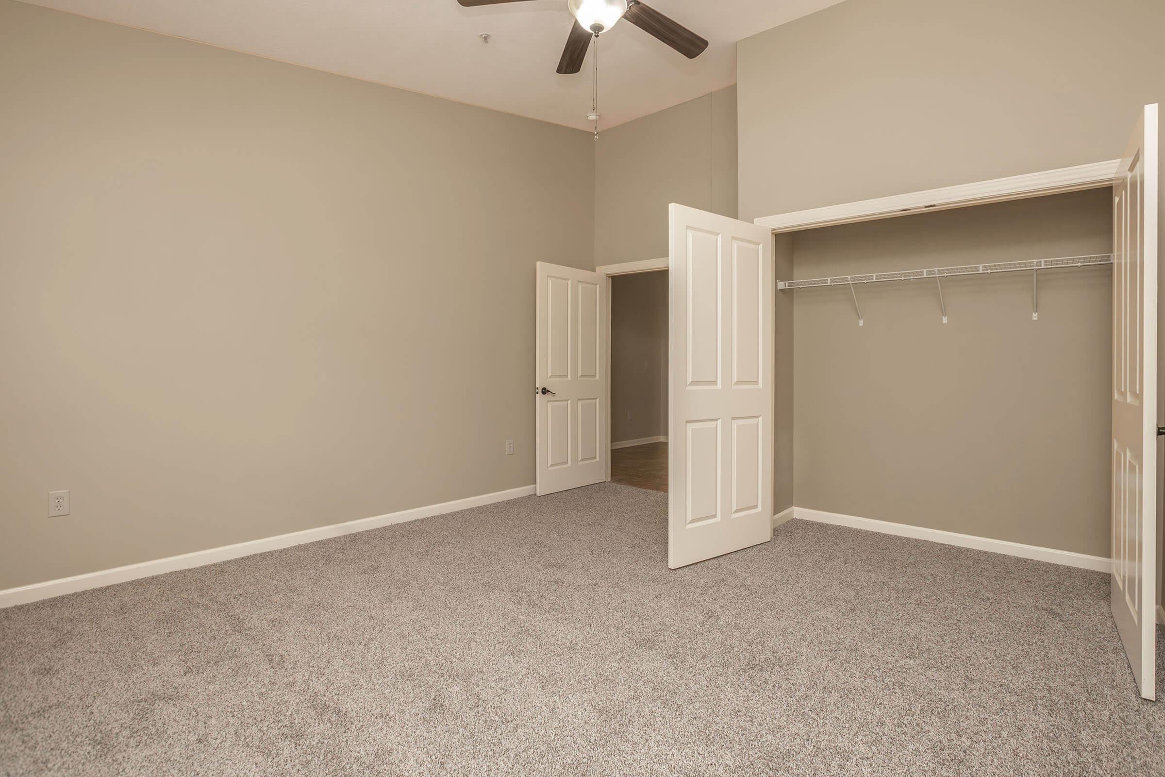A spacious room with light beige walls and carpeted flooring. It features a ceiling fan and two open doors leading to a closet area with a hanging rod. The room has a high ceiling, providing an airy feel, and is well-lit with natural light coming from an adjacent doorway.