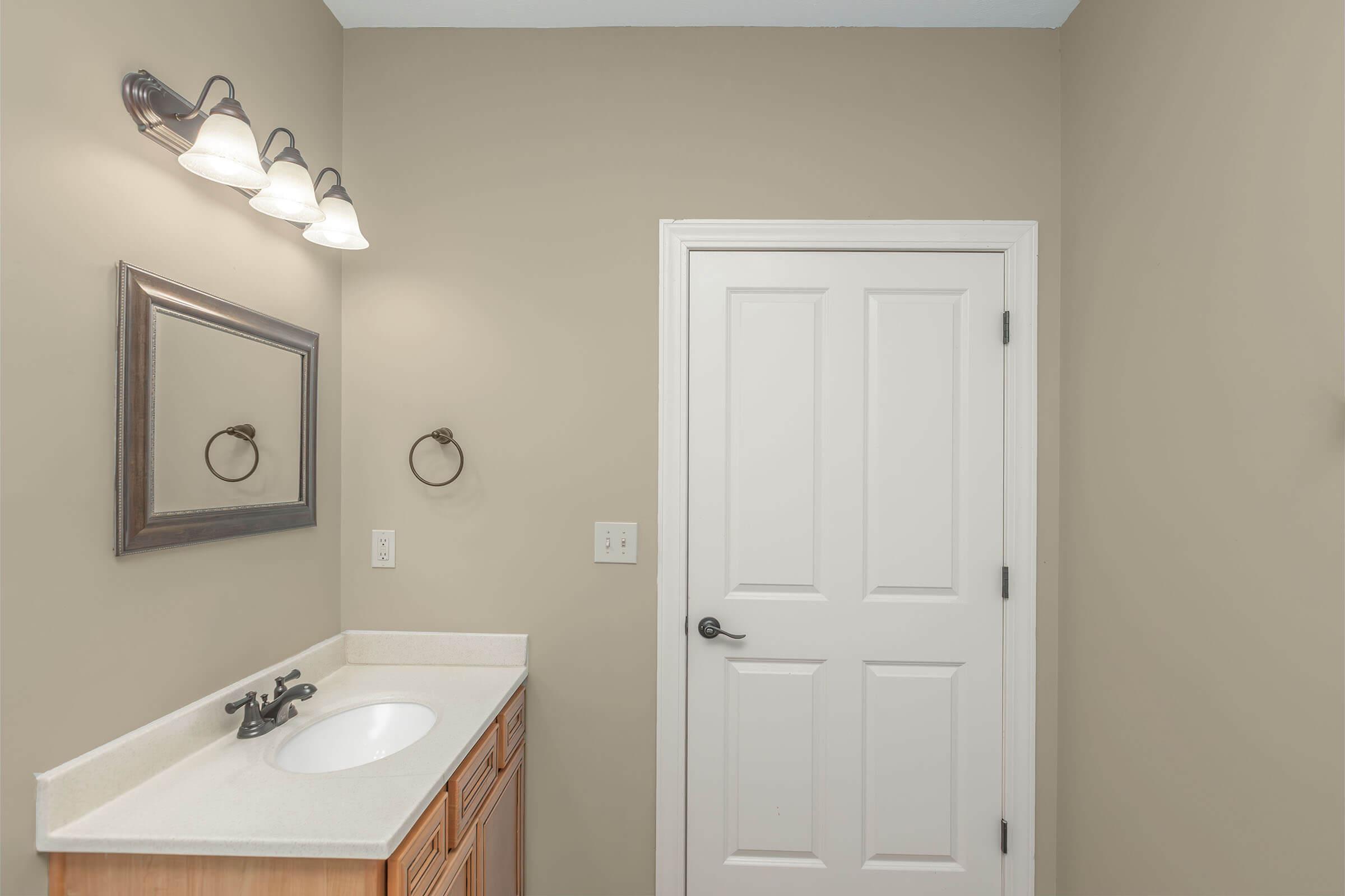 A well-lit bathroom featuring a single sink vanity with wooden cabinetry, a circular mirror above the sink, and a towel ring on the wall. The walls are painted a neutral color, and a white door is visible, leading to another area.
