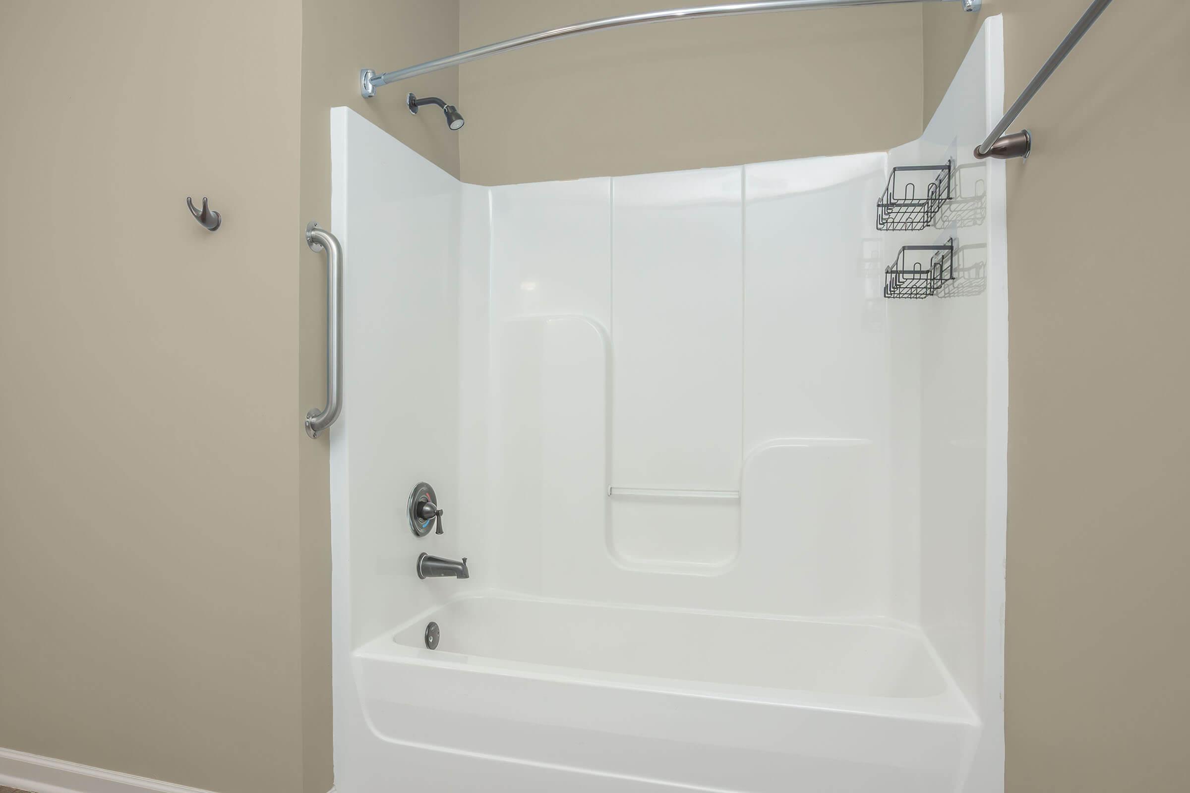 A clean, modern bathroom with a white shower and tub enclosure. The shower features a curved chrome rod and two wire storage caddies attached to the wall. The walls are painted a light beige, creating a bright and inviting atmosphere. A single hook is visible on the wall for towels or clothing.
