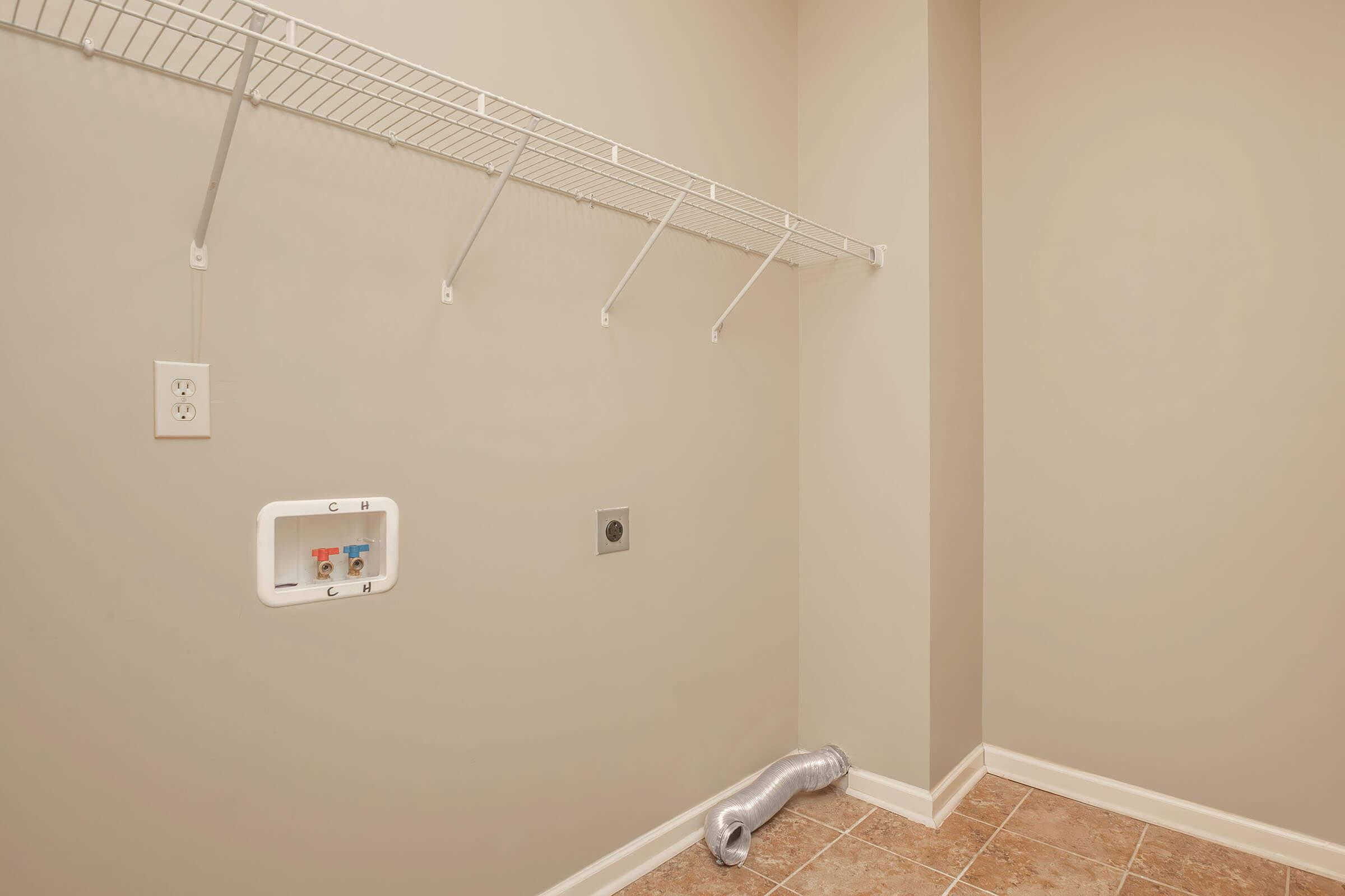 A clean, empty laundry room featuring a wall-mounted shelf with several hooks, an electrical outlet, and water connections. The floor is tiled, and there is a silver ducting hose on the floor, indicating it might be used for a dryer or ventilation system. The walls are painted a neutral color.