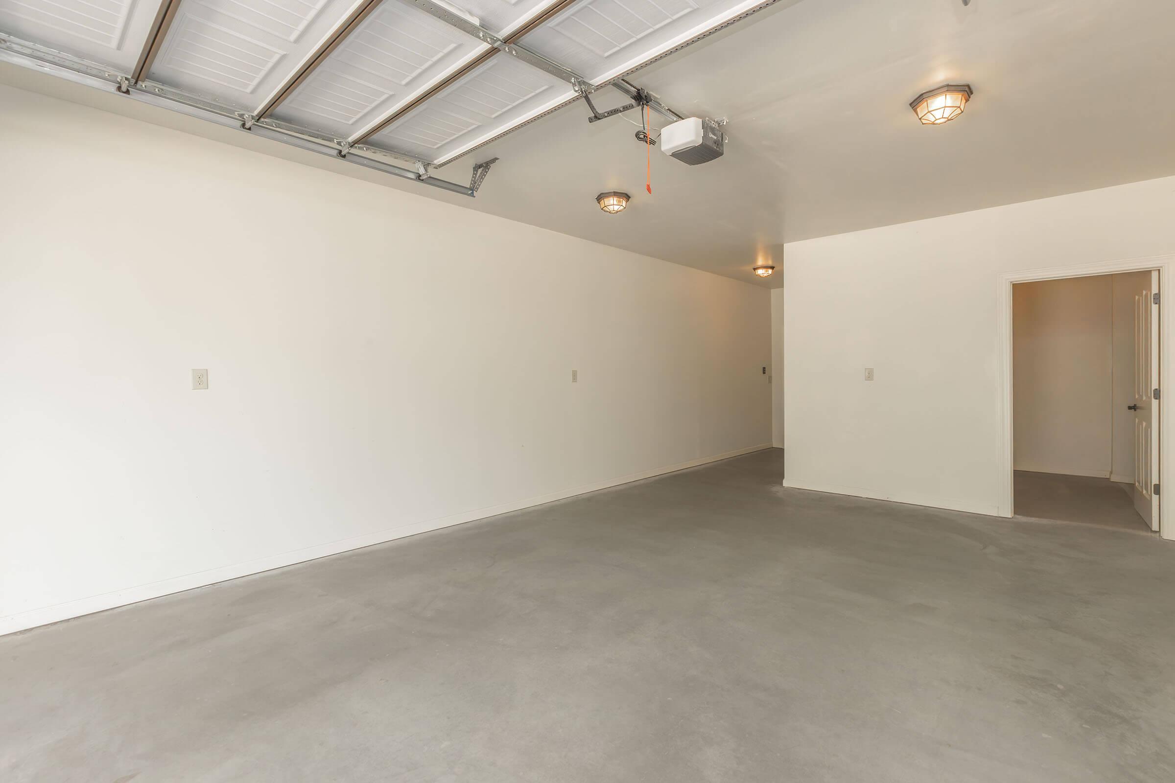 A spacious, empty garage with white walls, a concrete floor, and overhead lighting. There are two light fixtures on the ceiling and a door leading to another room on the right side. The garage has a clean, uncluttered appearance, ideal for storage or parking.
