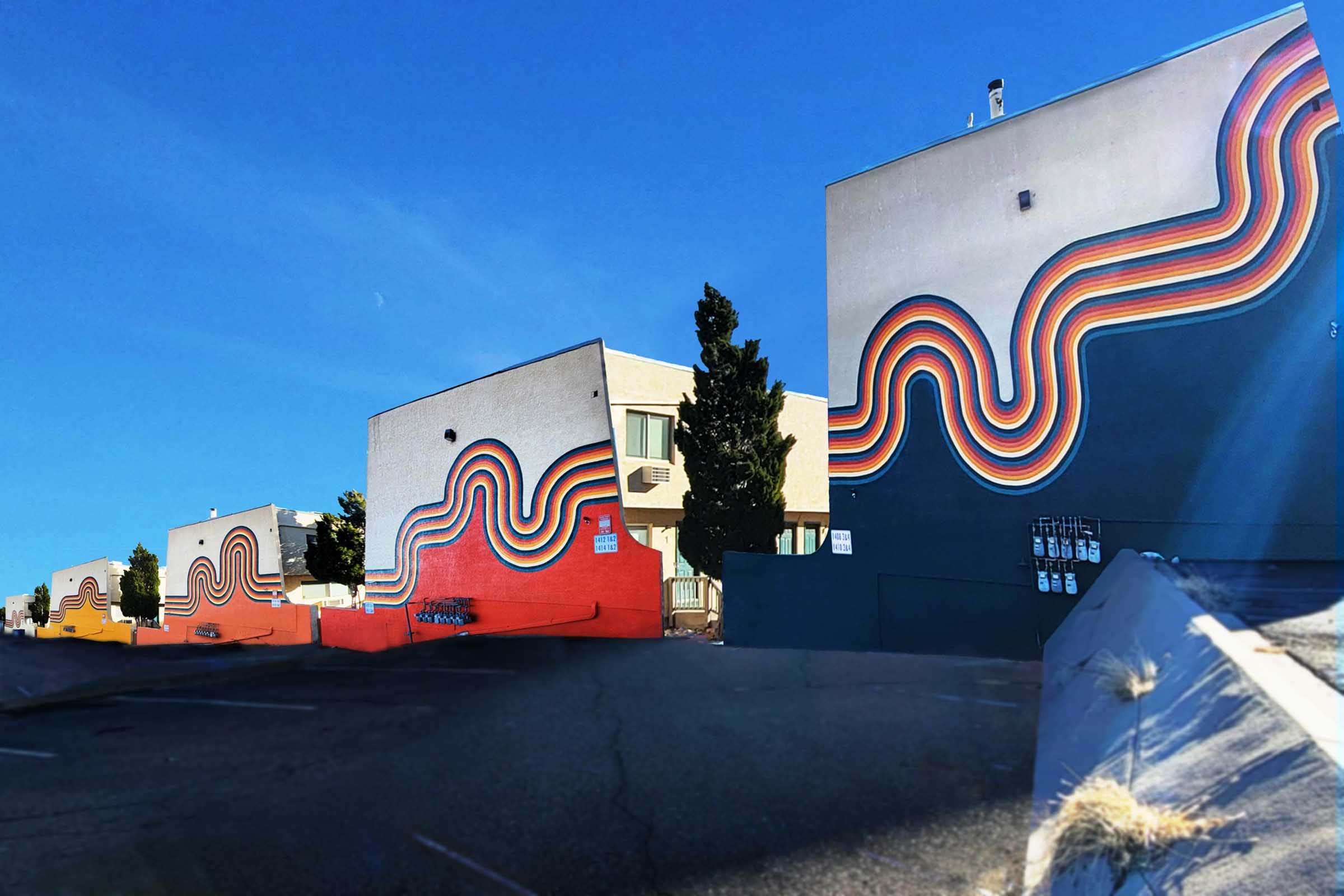 Colorful murals adorn the exterior walls of several buildings, featuring vibrant wavy lines in shades of red, orange, yellow, green, and blue. The clear blue sky and distant trees provide a bright backdrop, highlighting the artistic designs that create a lively urban atmosphere.