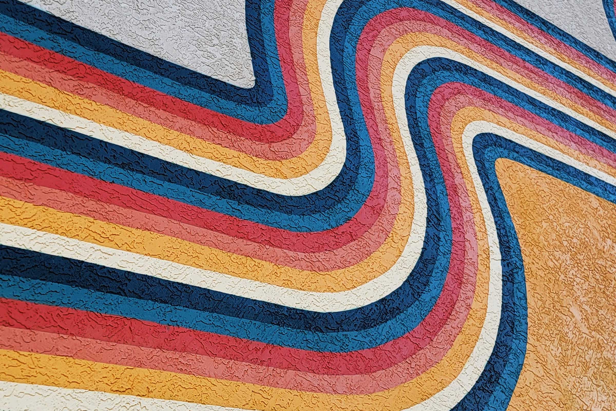 A vibrant abstract mural featuring a series of wavy stripes in colors including red, blue, yellow, and orange. The textured surface adds depth to the design, creating a dynamic visual effect. The pattern flows across the wall, evoking a sense of movement and energy.