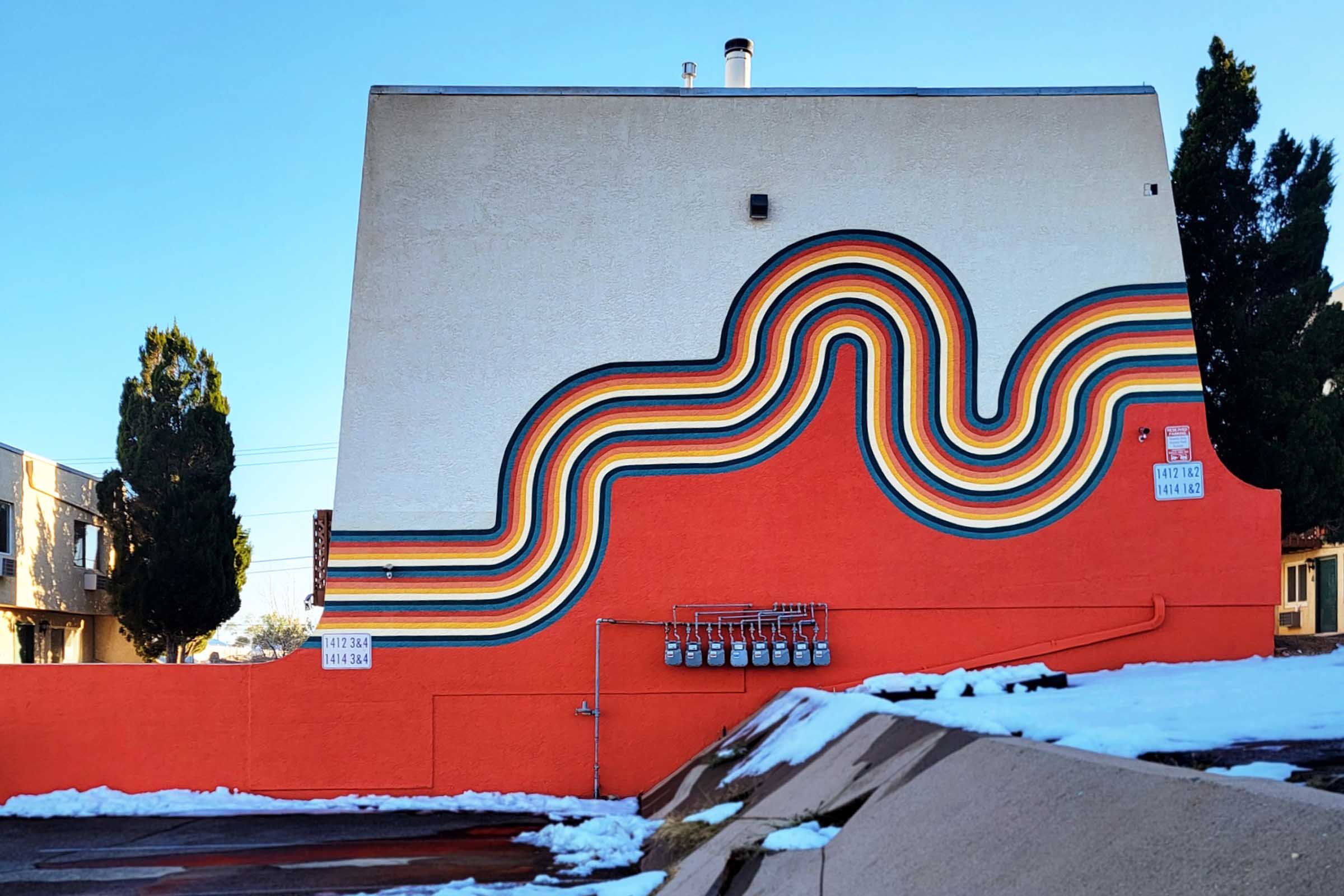 Colorful mural on the side of a building, featuring a wavy design in vibrant stripes of red, orange, yellow, green, and blue against a contrasting orange background. Utility meters are visible at the bottom, and snow is scattered on the ground in the foreground. The sky is clear and blue.