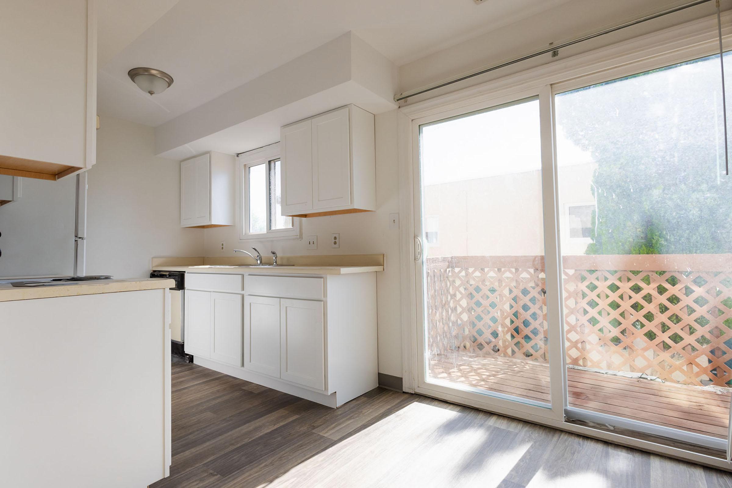 Bright and airy kitchen area featuring white cabinets, a countertop, and a sliding glass door that opens to a wooden deck. The space is well-lit with natural light coming through the windows, highlighting the clean and modern design. The flooring is light wood, adding warmth to the room.