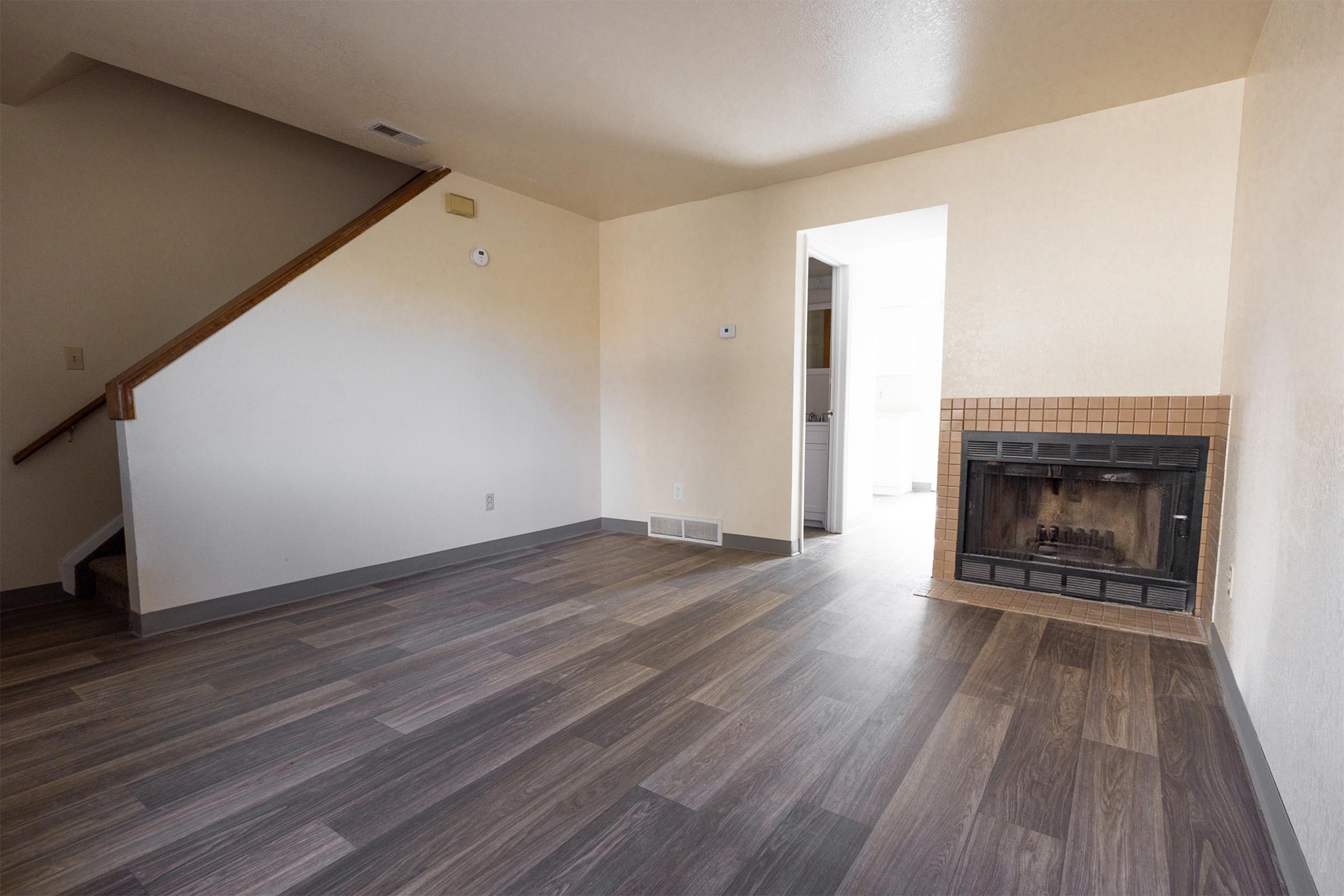 A spacious living room featuring a staircase on the left, a fireplace with a tiled surround, and light-colored walls. The flooring is a modern wood-like laminate, and there is a doorway leading to another room in the background, providing natural light.