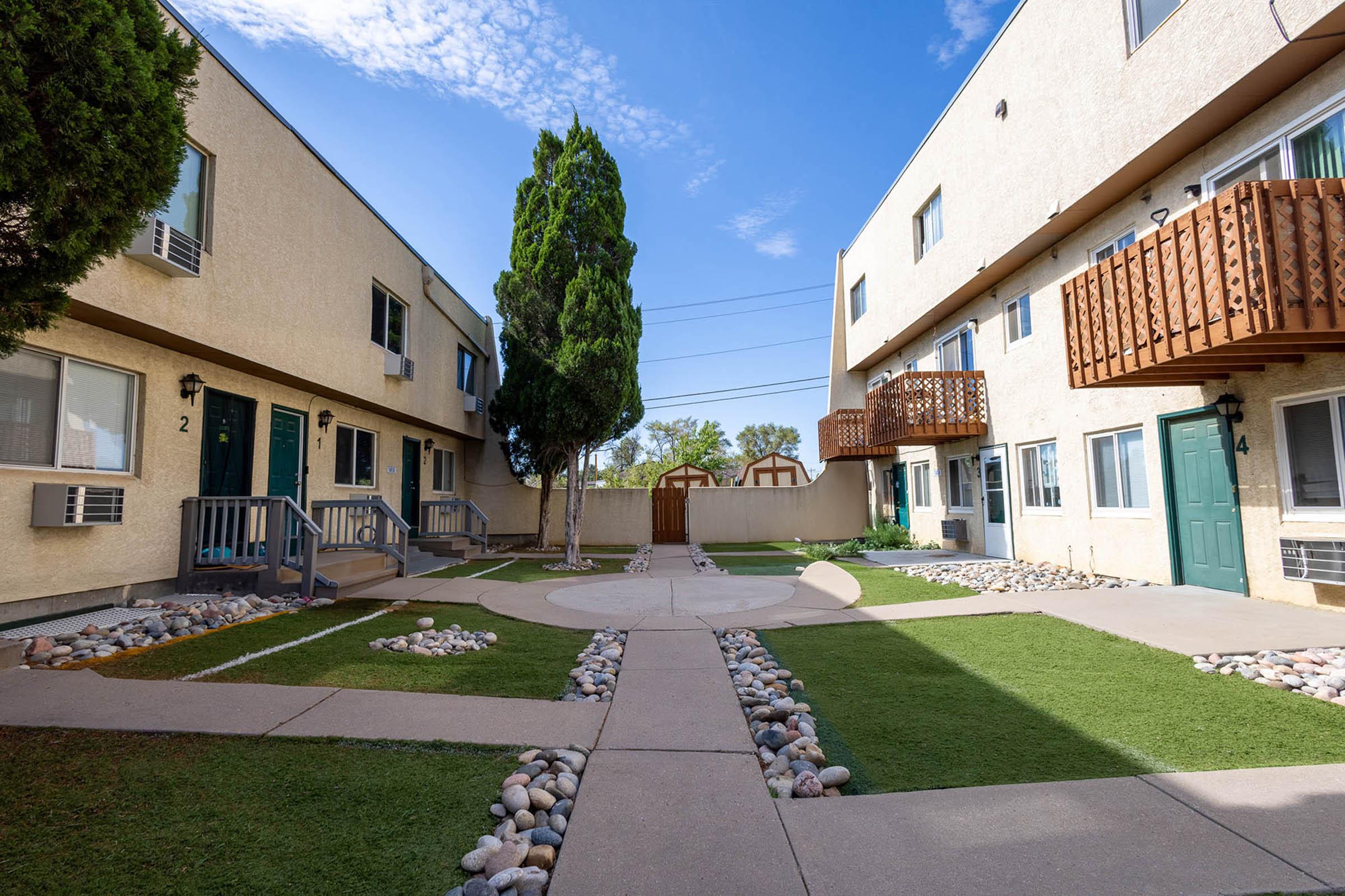 A small apartment complex featuring two-story buildings with balconies and air conditioning units. The courtyard includes neatly laid pathways, grass, and decorative stones, surrounded by trees. Bright blue skies are visible, enhancing the outdoor ambiance.