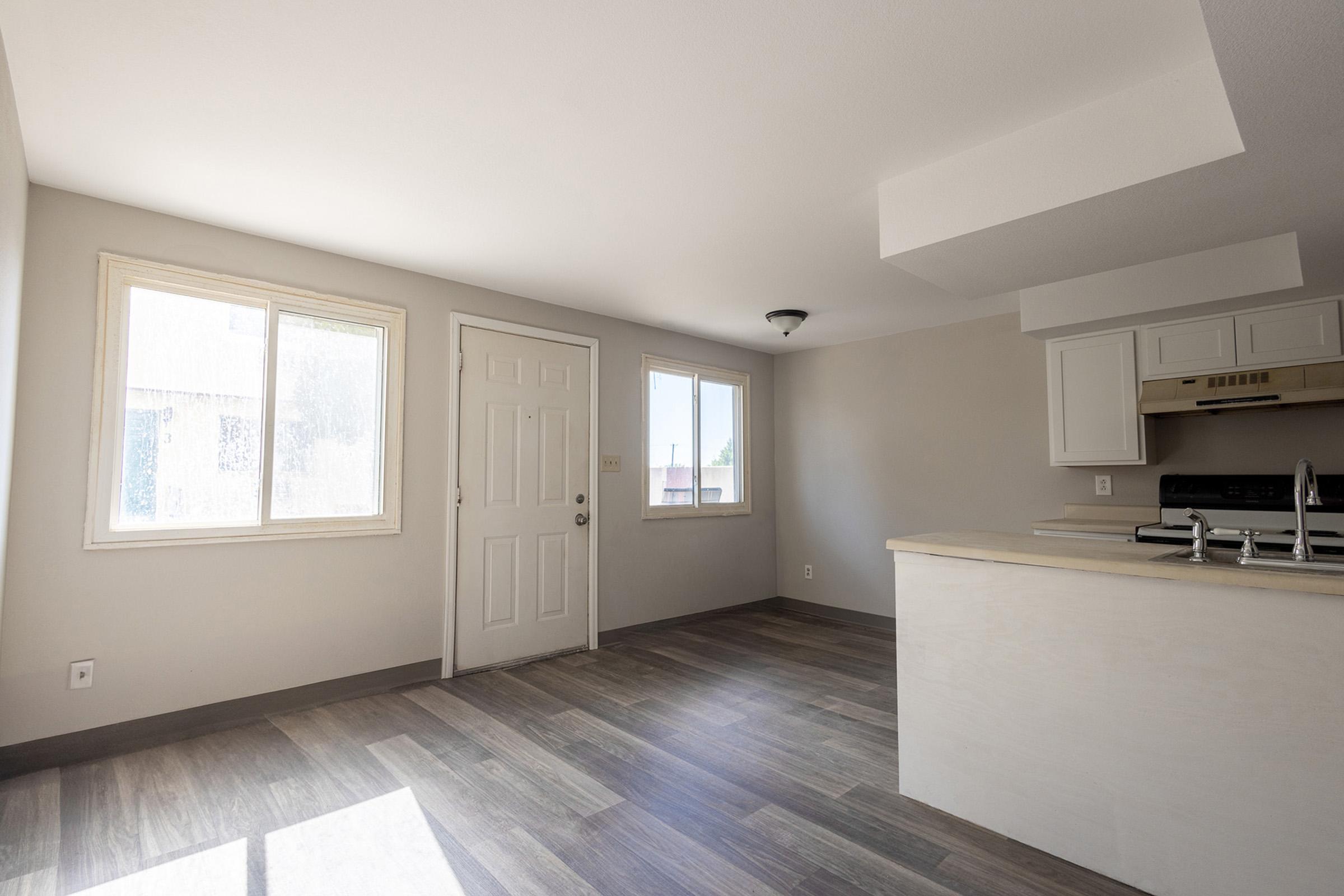 A spacious, empty room featuring light-colored walls and a modern kitchen area with white cabinetry. Large windows allow natural light to fill the space, creating a bright atmosphere. The flooring is made of dark wood laminate, enhancing the contemporary feel of the area.