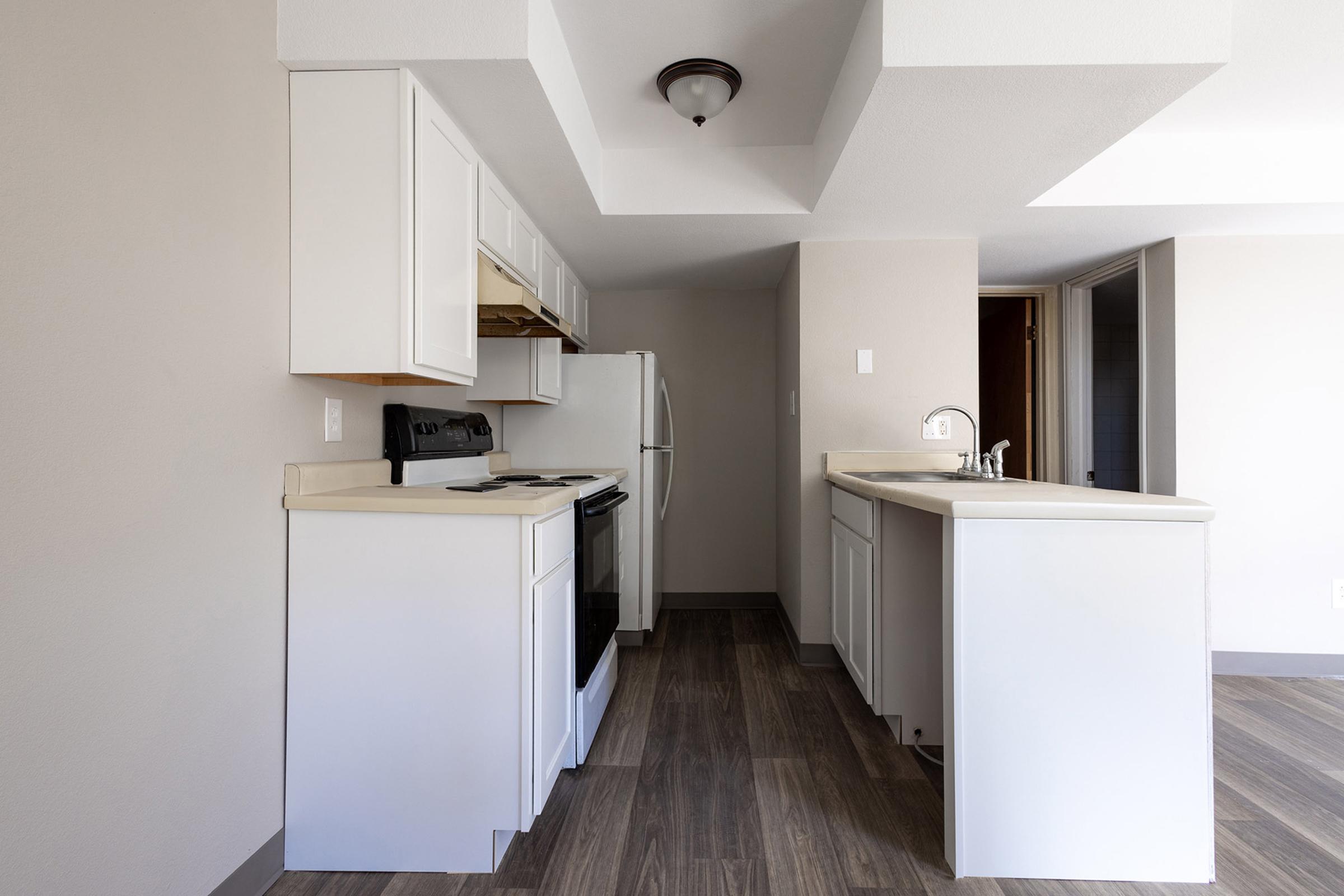 Modern kitchen with white cabinetry, a stovetop, and a refrigerator. The countertop is light-colored, and the flooring is wood laminate. The space is open, featuring a small island with seating. Natural light comes from the adjacent room, highlighting the streamlined design and neutral color palette.