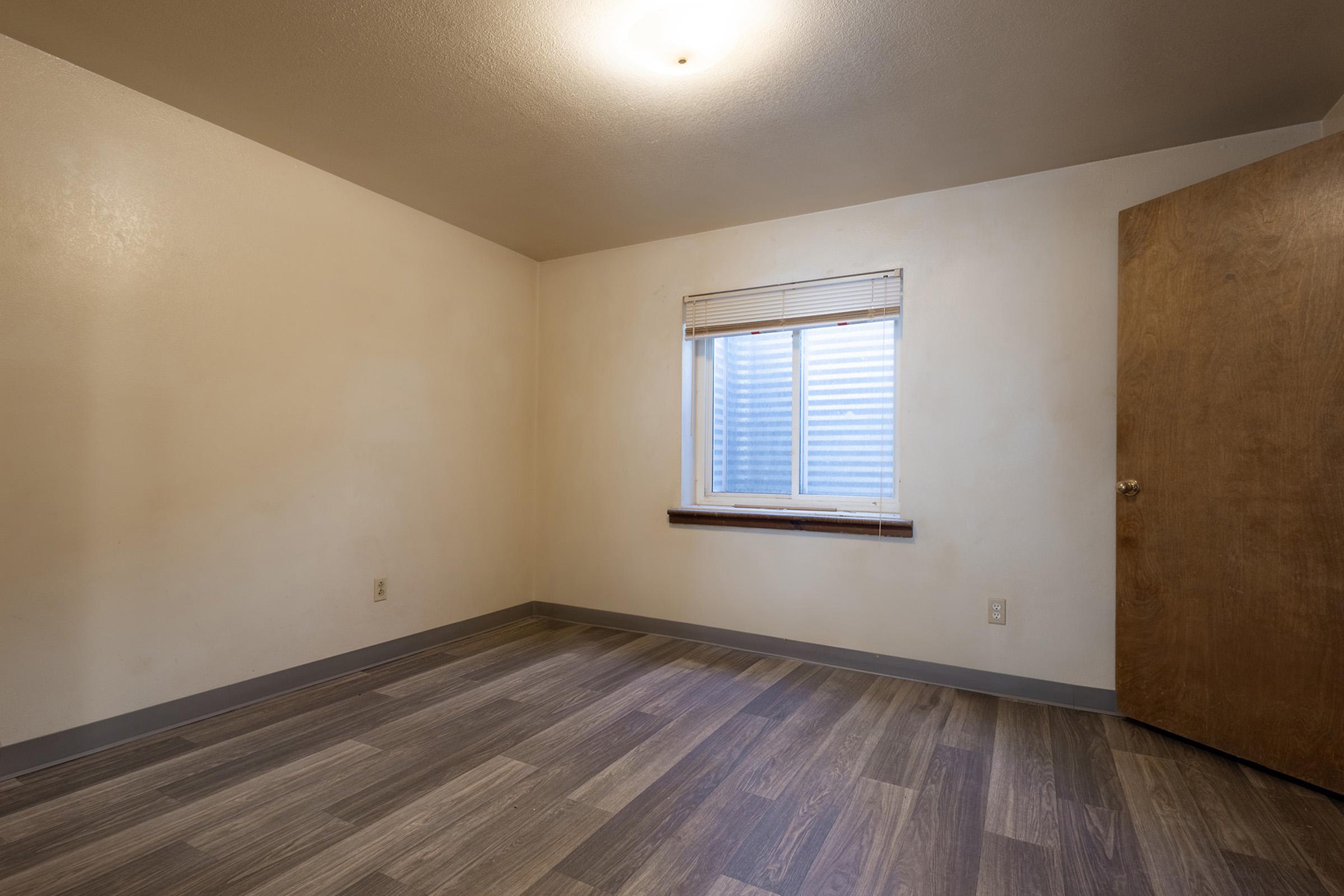 Empty interior room with light-colored walls, a window with blinds allowing natural light, and a wooden door. The floor features laminate wood planks, and there are no furnishings present, creating a clean and spacious atmosphere.