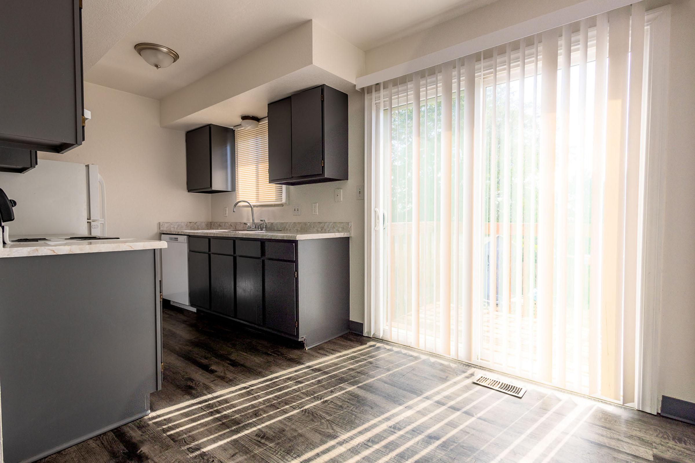 A modern kitchen featuring dark cabinets and a spacious layout. Light streams in through vertical blinds on a glass door, illuminating the room with warm tones. The countertop is a light marble pattern, and the floor has a sleek, dark finish, enhancing the contemporary feel of the space.