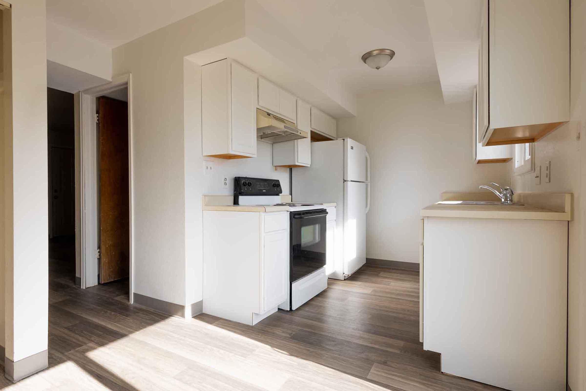 A modern kitchen featuring white cabinetry, a stove, refrigerator, and sink against a light-colored wall. The floor is made of wood-like material, and there is natural light coming from a nearby window, creating a bright and airy atmosphere.