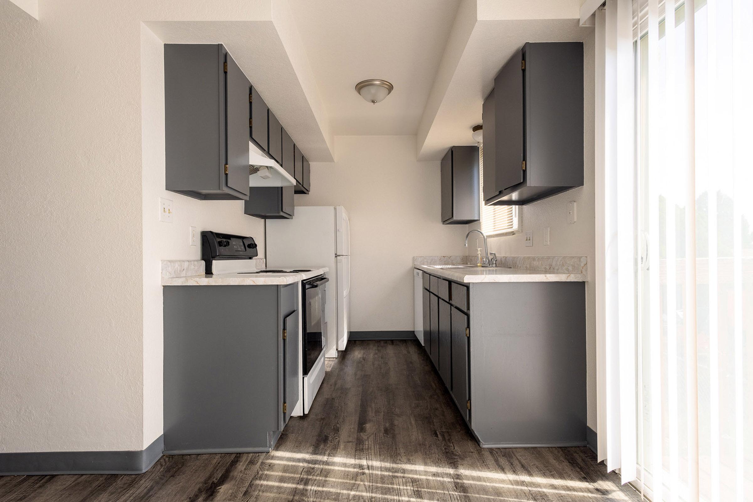 A modern kitchen featuring gray cabinets, a white countertop, an electric stove, and a refrigerator. The space includes a sink with a faucet and has large windows with vertical blinds letting in natural light. The flooring is a dark wood laminate, creating a warm and inviting atmosphere.