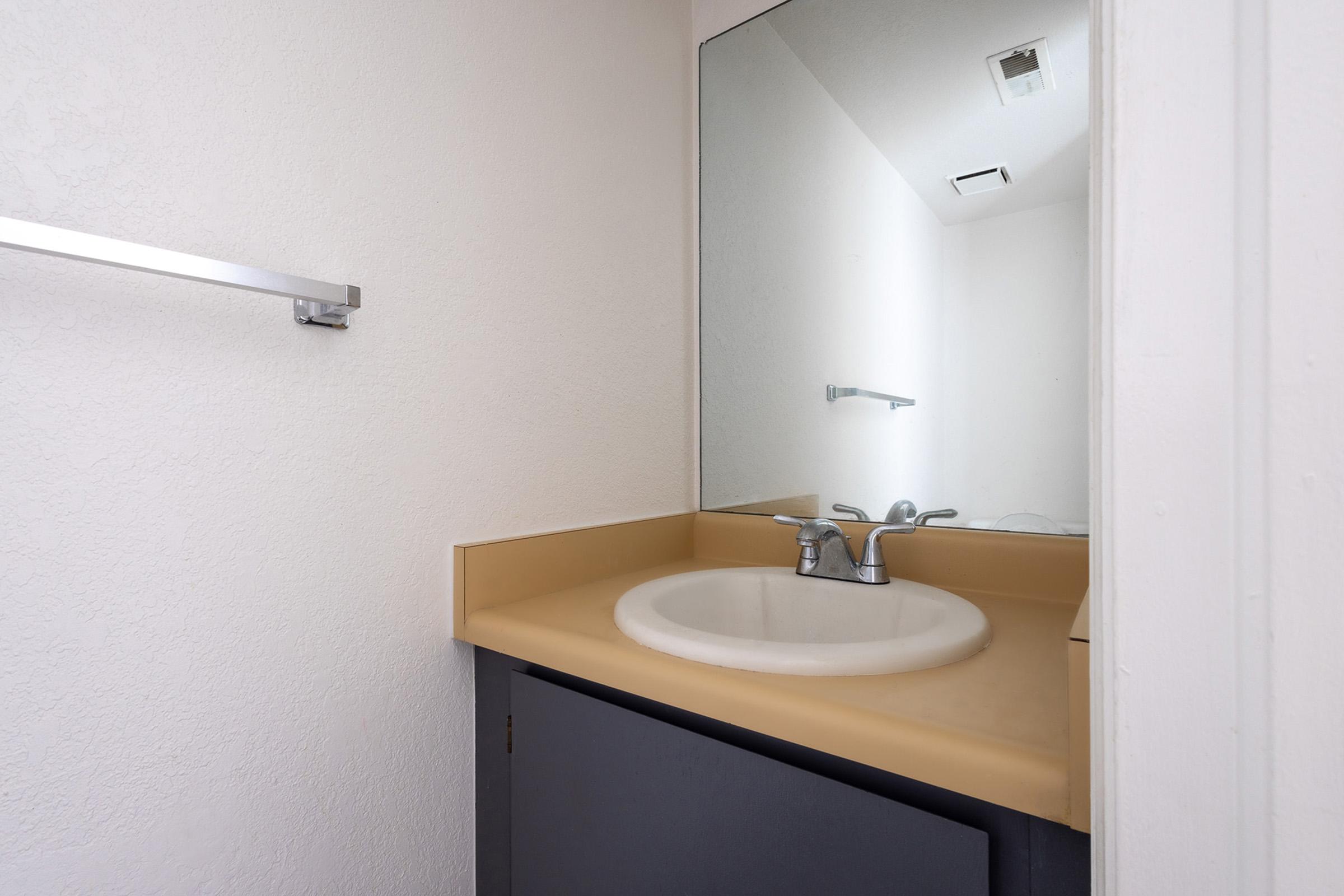 A small bathroom with a simple countertop and sink, featuring a single faucet. The wall is painted white, and there's a mirror above the sink. A towel rack is mounted on the wall, and the cabinetry below the sink is dark-colored, providing a clean and minimalist aesthetic.