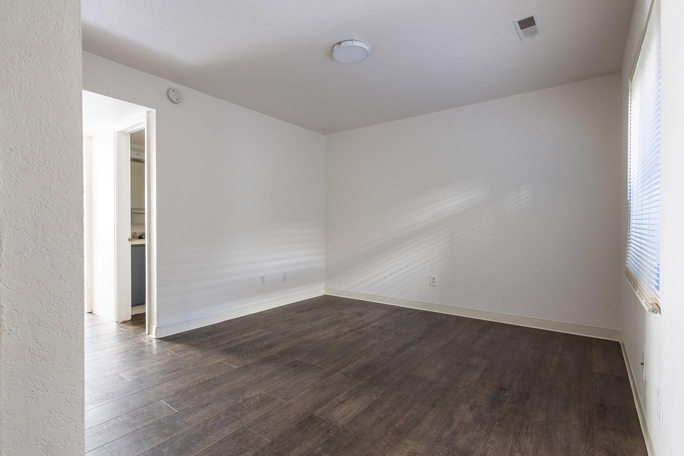 An empty room with wooden flooring, light-colored walls, and a ceiling light fixture. A doorway leads to another room on the left. Natural light enters through a window, casting soft shadows across the floor. The space is uncluttered and waiting for furniture or decoration.