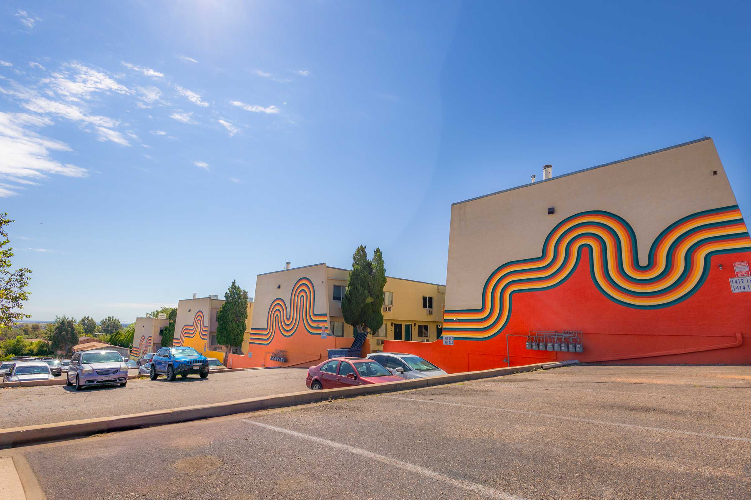 Colorful murals featuring wavy rainbow patterns adorn the exterior of several buildings. The scene takes place on a sunny day with a clear blue sky. Below, a parking lot is visible with a few cars parked. Trees are present alongside the buildings, adding greenery to the vibrant atmosphere.