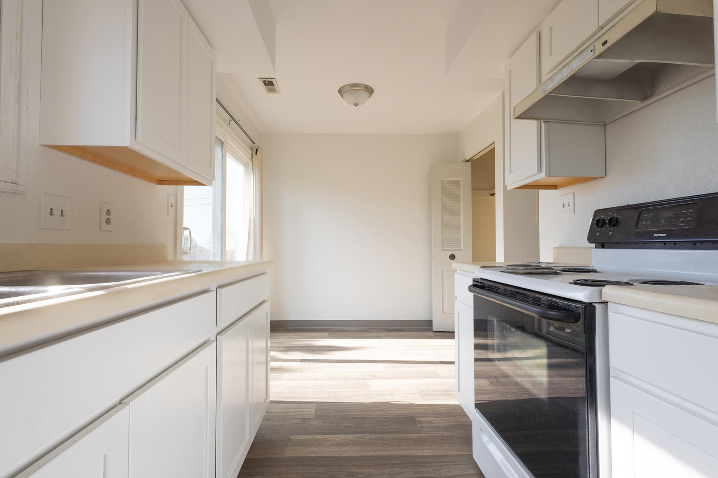 A bright, modern kitchen featuring white cabinetry, a black stove, and a countertop with a sink. Natural light floods in through a window, emphasizing the spaciousness and clean design. The floor is covered in light wood-like tiles, creating an inviting atmosphere.