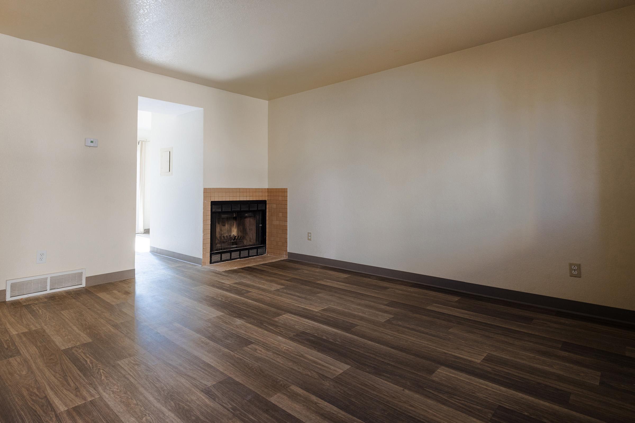 Interior of a spacious living room with light-colored walls and a hardwood floor. A fireplace is visible on the left side, and a doorway leads to another room, suggesting natural light coming from the adjacent space. The overall ambiance is bright and inviting, with minimal furnishings.