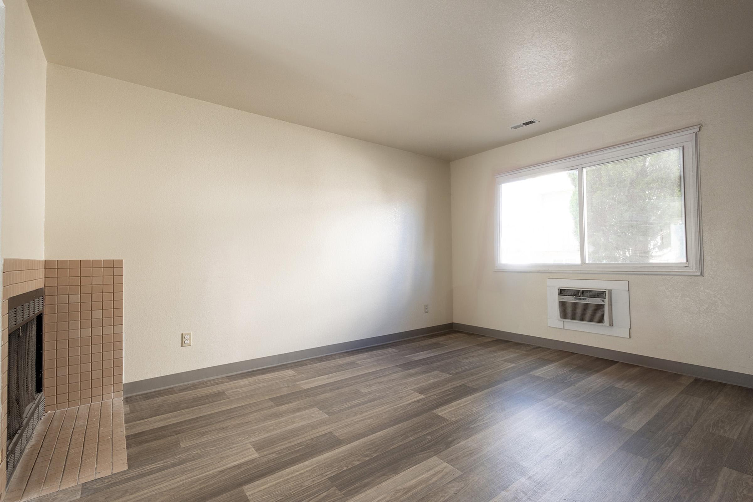 A spacious, empty room with light-colored walls and a window letting in natural light. The floor is covered with wooden-looking laminate. There's a corner fireplace with a pink tile surround, and an air conditioning unit installed on the wall below the window.