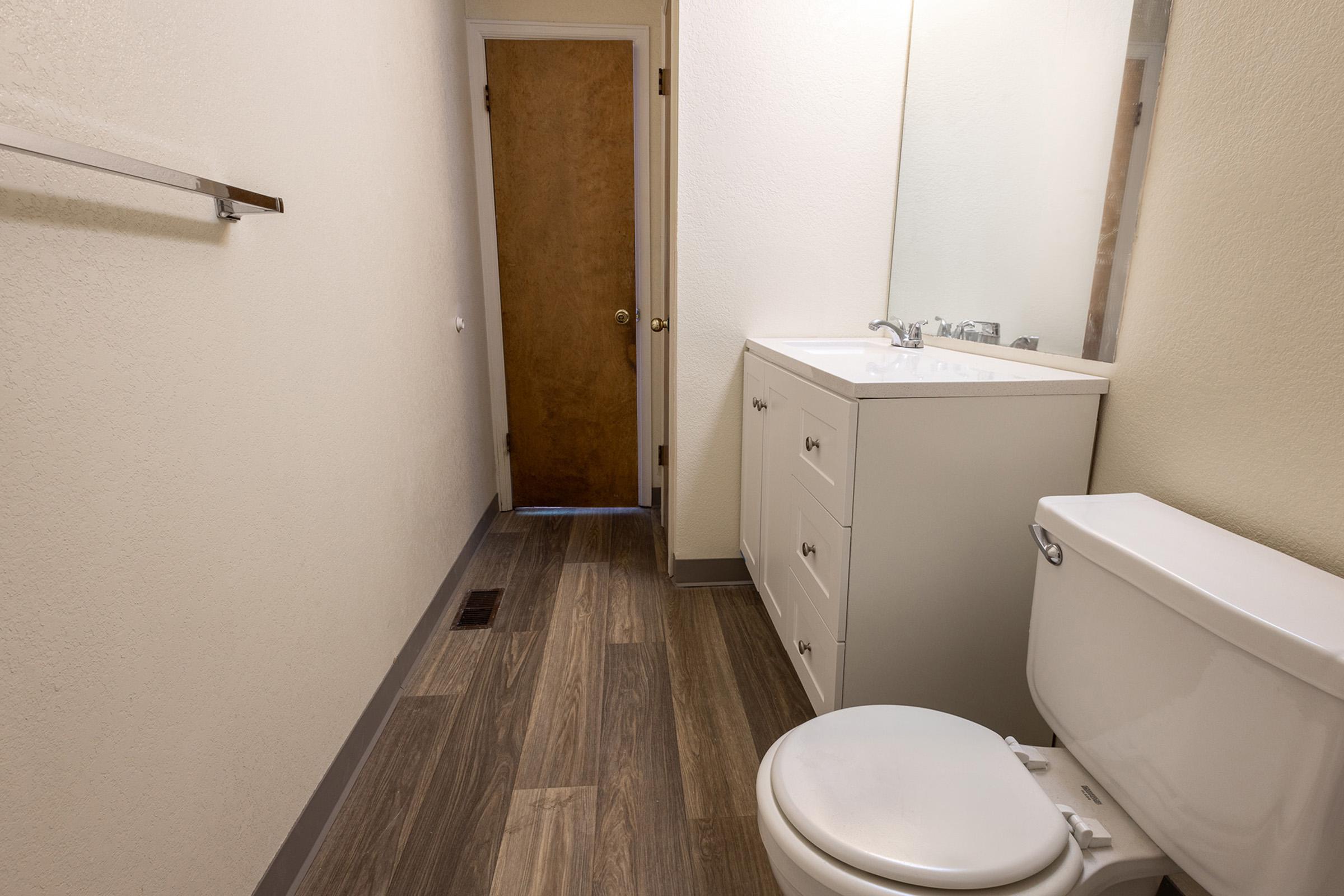 A clean, modern bathroom featuring a white vanity with drawers, a toilet, and a large mirror. The walls are painted light, and the floor is covered with wood-like tiles. There's a wooden door at the end of the hallway, and a towel rack mounted on the wall. The space is well-lit and organized.