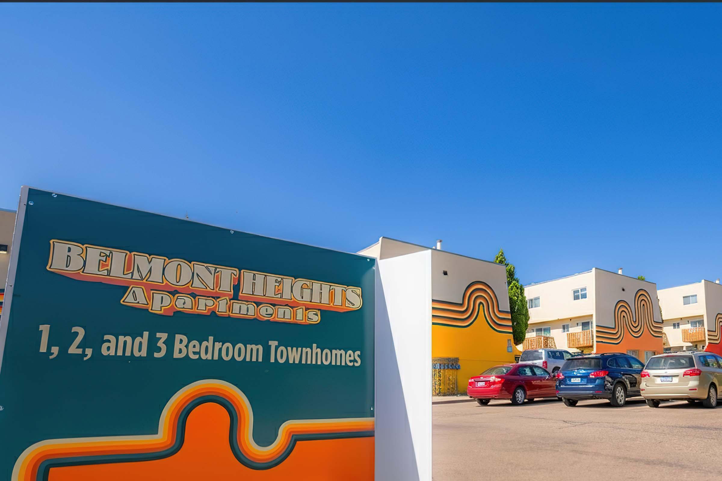 A colorful sign for Belmont Heights Apartments with text indicating 1, 2, and 3-bedroom townhomes. The background features modern apartment buildings with vibrant designs and multiple parked cars under a clear blue sky.