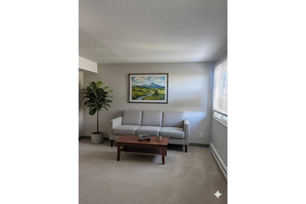 A cozy living room featuring a light grey couch with a wooden coffee table in front. A potted plant is beside the couch, and a landscape painting hangs on the wall. Natural light streams in from a window, illuminating the neutral-toned decor and carpet.