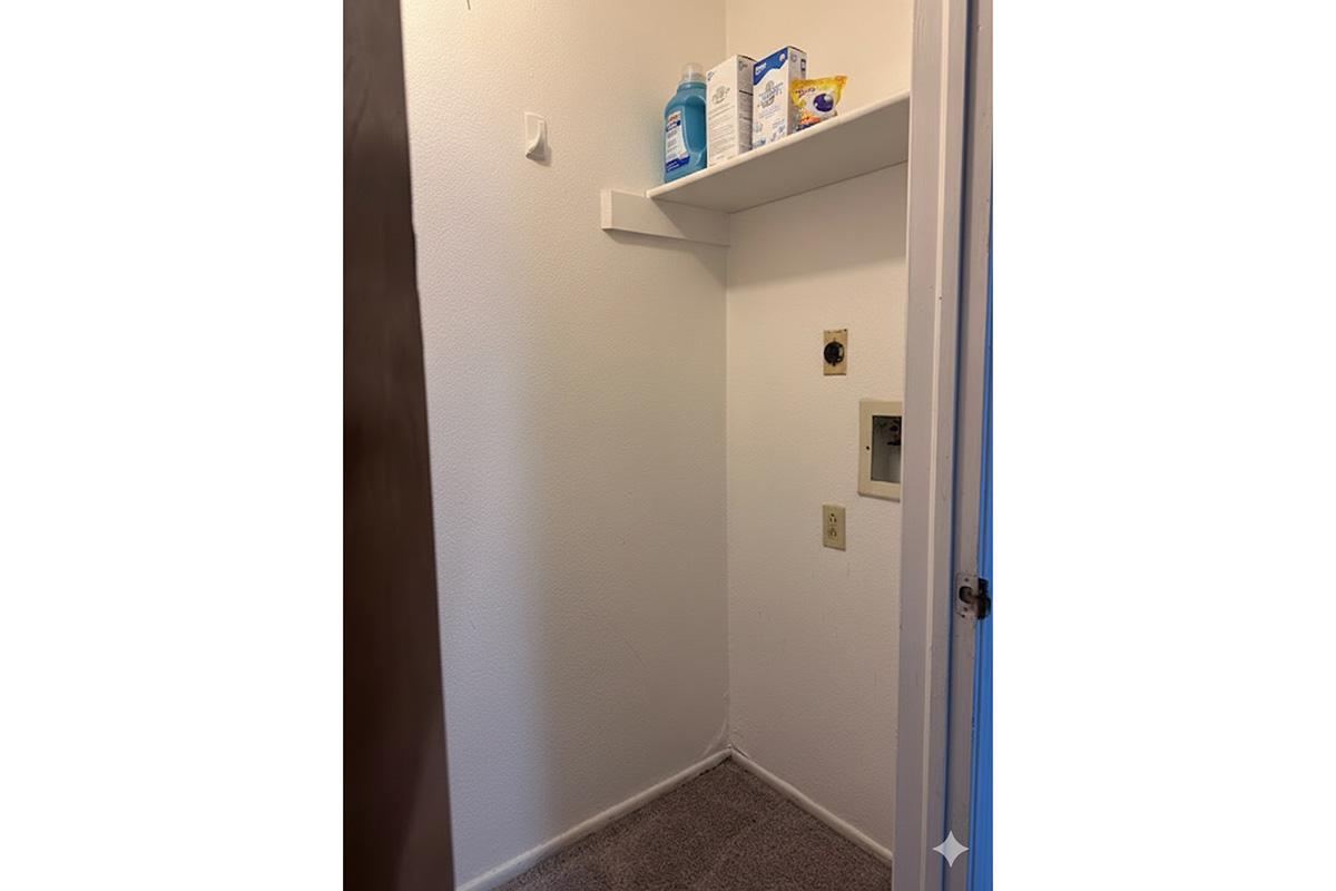 Inside a small, empty corner room with white walls and gray carpet. A single shelf holds cleaning supplies, and there are a few electrical outlets on the wall. The space is sparsely furnished with a door partially open at the edge of the image.