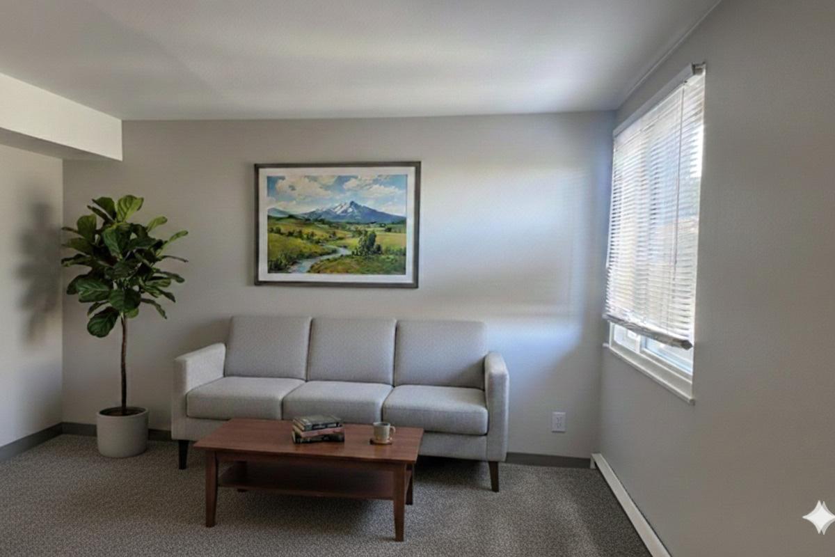 A cozy living room featuring a light grey sofa, a wooden coffee table with a book on it, and a tall green plant. A large painting of a mountainous landscape hangs on the wall, and natural light streams in through a window with white blinds, enhancing the warm, inviting atmosphere.
