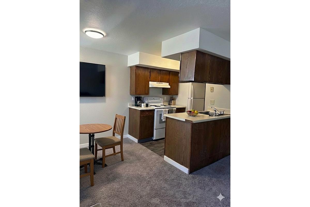 A cozy kitchen and dining area featuring a small wooden table with two chairs, a compact kitchen with brown cabinets, a white stove, and a refrigerator. There is a flat-screen TV mounted on the wall, and the floor is covered with light carpeting. The space is well-lit with soft overhead lighting.