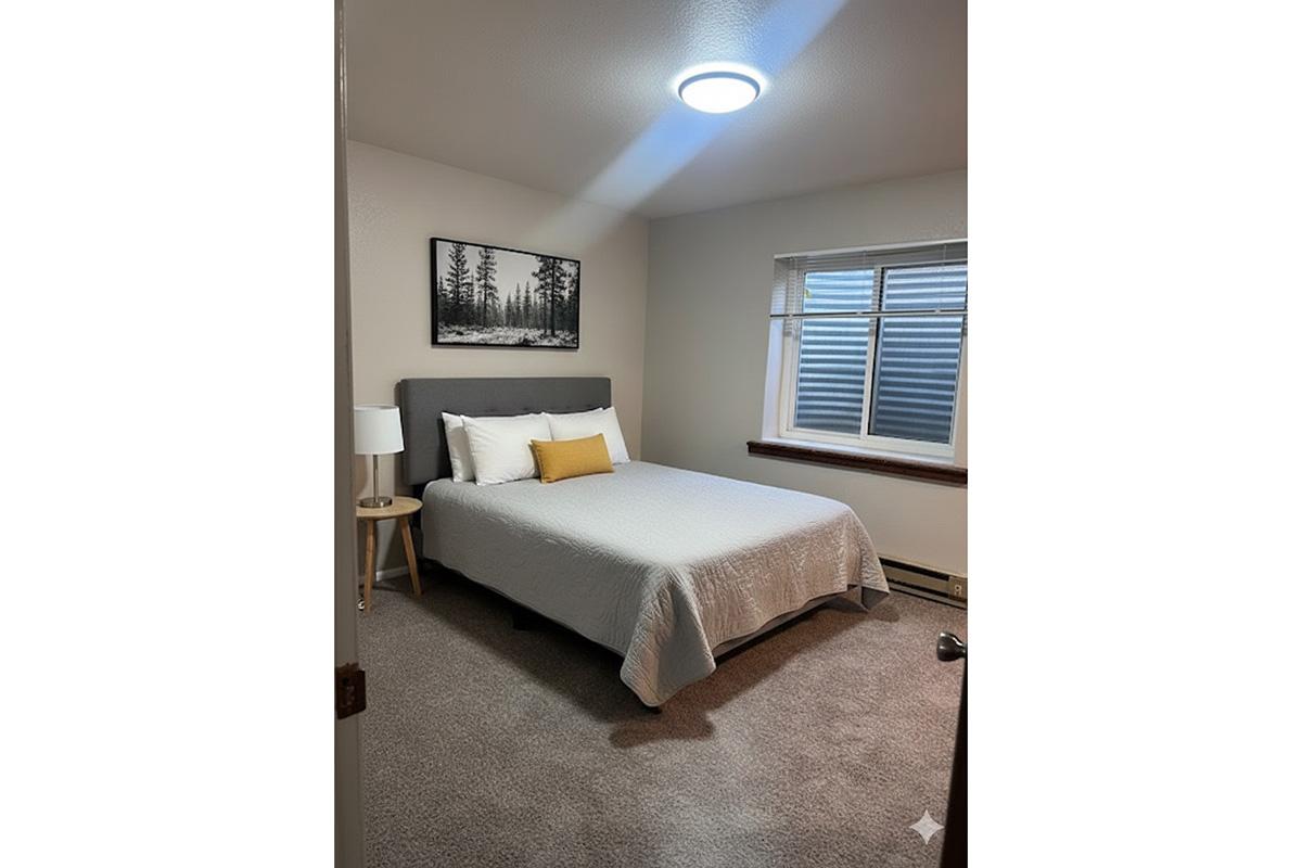 A cozy bedroom featuring a queen-sized bed with a gray upholstered headboard, white bedding, and a yellow decorative pillow. There's a wooden nightstand with a lamp and a forest-themed artwork on the wall. A window allows natural light, and the floor is covered with light-colored carpet.