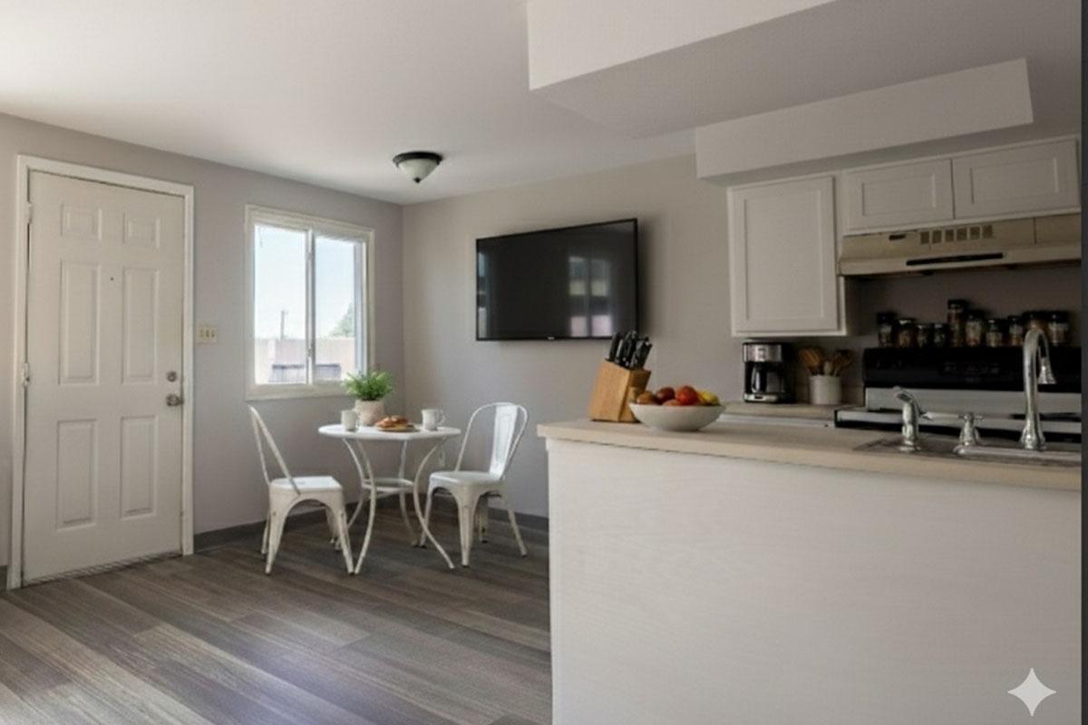 A modern kitchen and dining area featuring a small table with two chairs, a doorway to the outside, and a window allowing natural light. There is a flat-screen TV mounted on the wall, and the kitchen includes a stove, sink, and counter space with a knife block and fruit bowl. The flooring is light-colored.