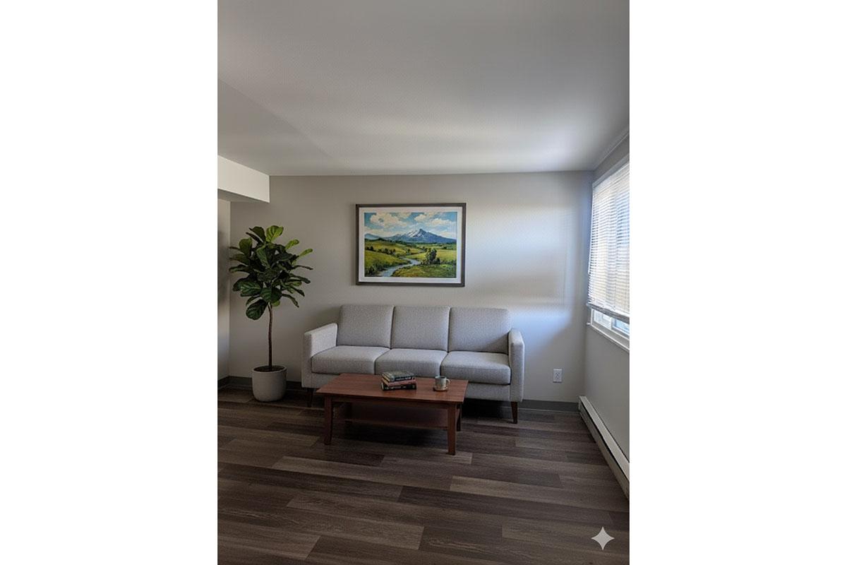 A modern living room featuring a light gray sofa with a wooden coffee table. A potted plant stands beside the sofa. There is a scenic landscape painting on the wall above the sofa, and natural light comes through the window, illuminating the space. The flooring is a warm-toned wood.