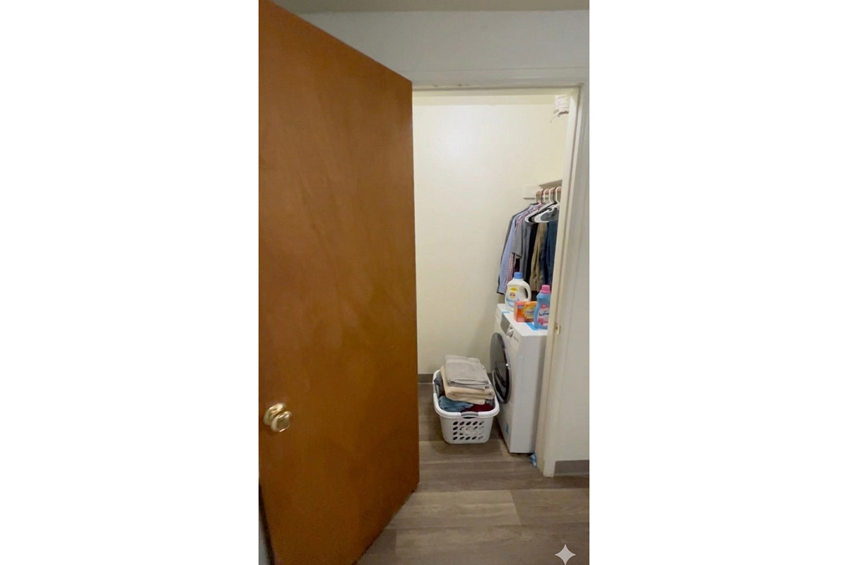 A partially open door leading to a small laundry area. Inside, there is a washing machine, a laundry basket filled with clothes, and clothes hanging on a rack. The floor is wooden, and the walls are light-colored.