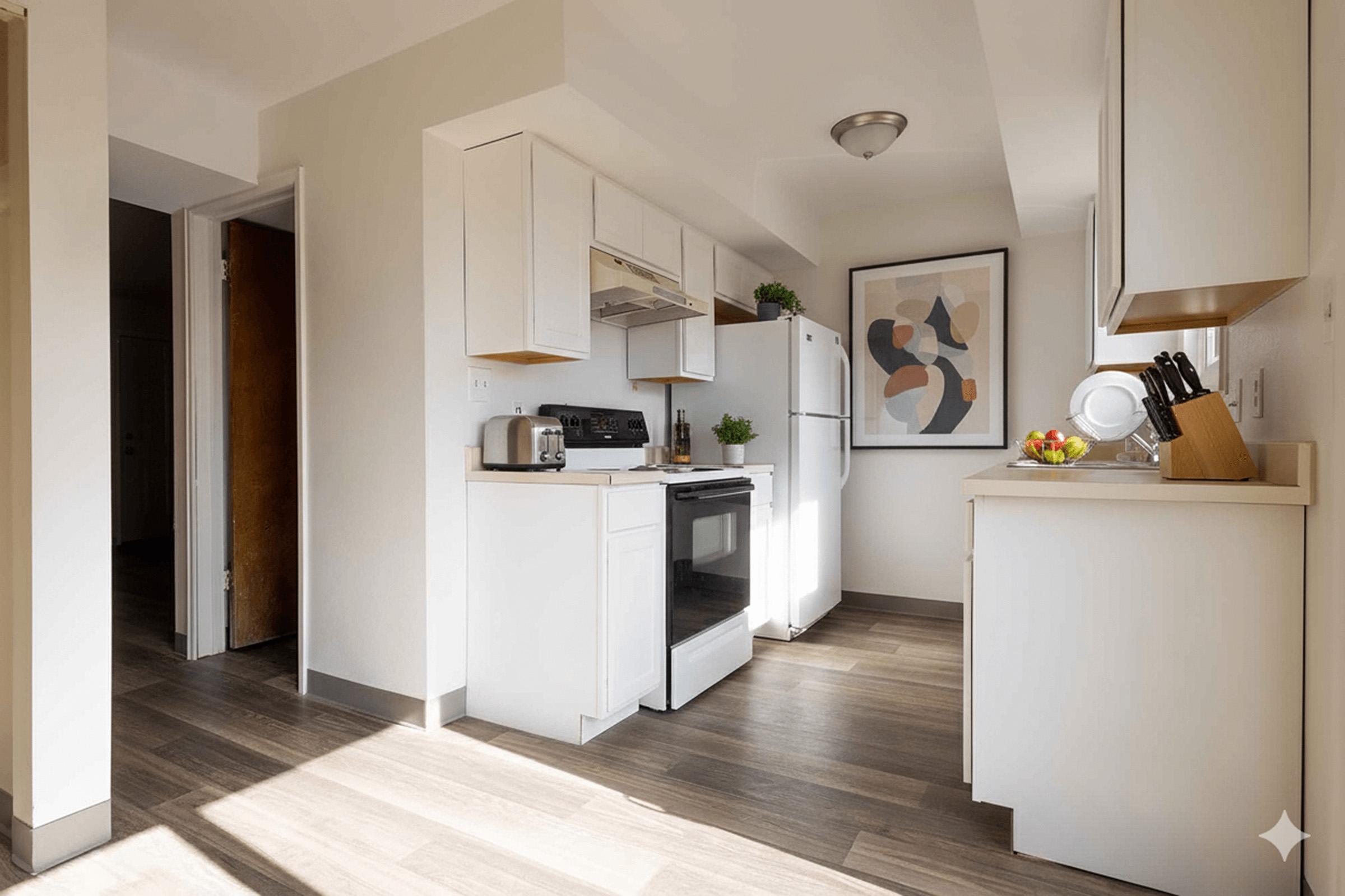 A modern kitchen featuring white cabinets and appliances, including a stove and refrigerator. The room has natural light streaming in, highlighting the wooden floor. A decorative artwork is displayed on the wall, and a bowl of fruit is on the counter. The design is clean and minimalistic.