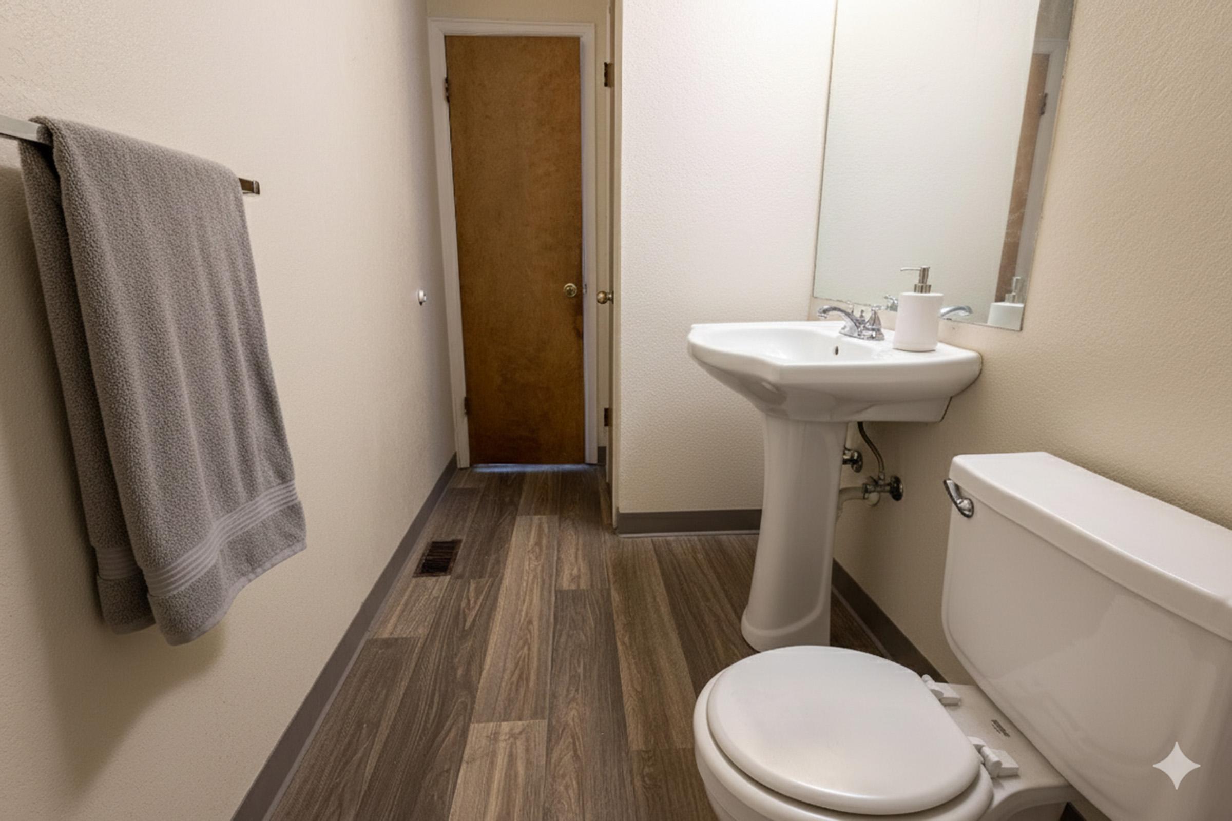 A clean, neutral-colored bathroom featuring a white toilet, a pedestal sink with a mirror above it, and a towel hanging on a rack. The floor has wooden tiles, and there's a brown door visible in the background, indicating a well-maintained and functional space.
