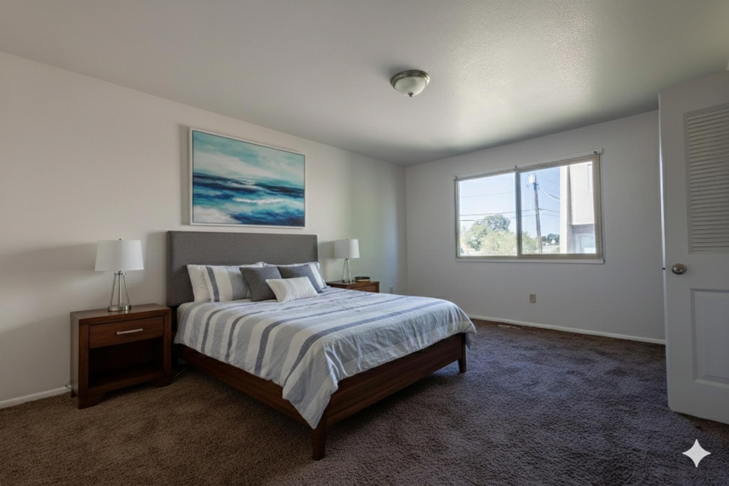 A bright and spacious bedroom featuring a large bed with striped bedding, two bedside lamps on wooden nightstands, and a piece of abstract wall art. Natural light comes through a window, illuminating the room, which has a neutral color scheme and plush carpet.