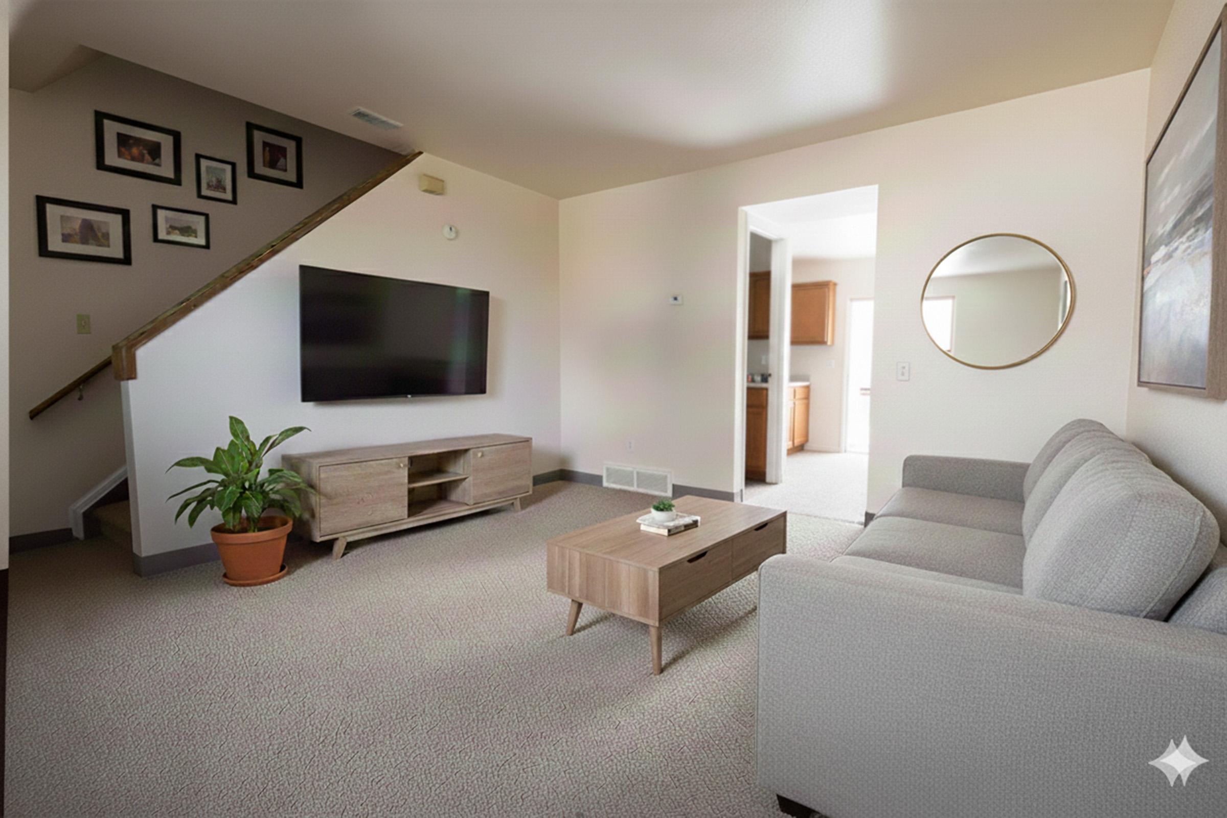 A modern living room featuring a gray sofa and a wooden coffee table on a carpeted floor. A large television is mounted on the wall, and a round mirror is located nearby. A potted plant adds a touch of greenery. Stairs lead to an upper level, and a doorway opens into a kitchen area in the background.