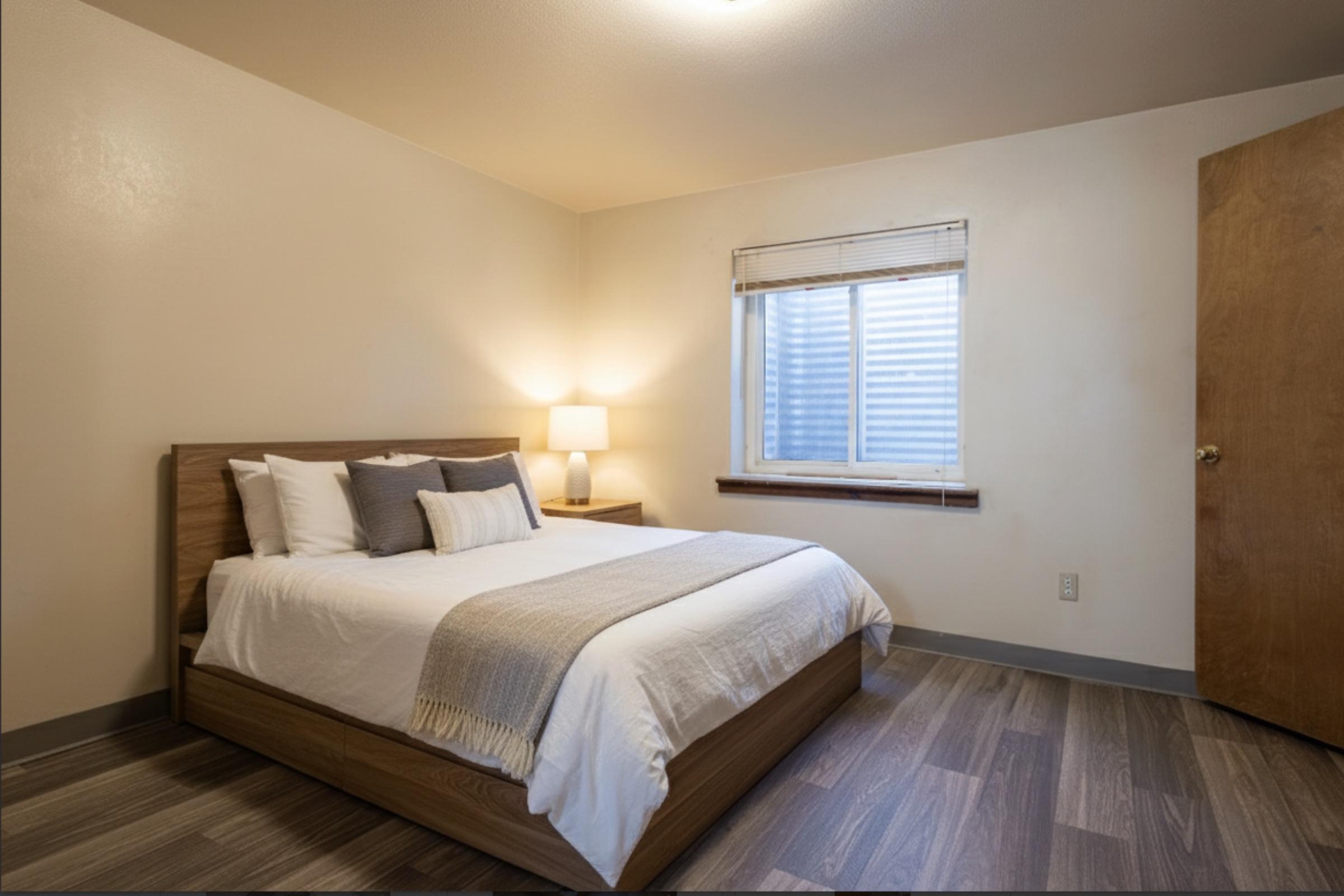 A cozy bedroom featuring a wooden bed with white linens and decorative pillows. Next to the bed is a bedside lamp on a small table. A window with sheer curtains allows natural light to brighten the room, which has wooden flooring and neutral-colored walls. A wooden door is visible on the right side.