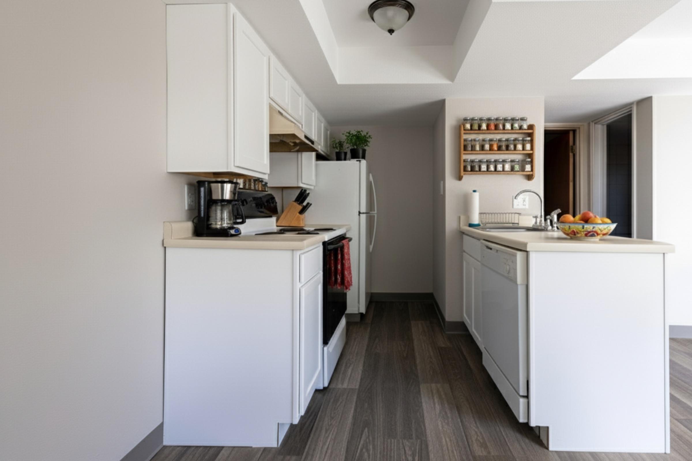 Modern kitchen with white cabinetry, countertop, and appliances. It features a coffee maker, cutting board, and a bowl of fruit on the counter. A spice rack hangs on the wall, and the flooring is a warm wood tone, enhancing the bright and welcoming atmosphere. Natural light streams in from an adjacent room.
